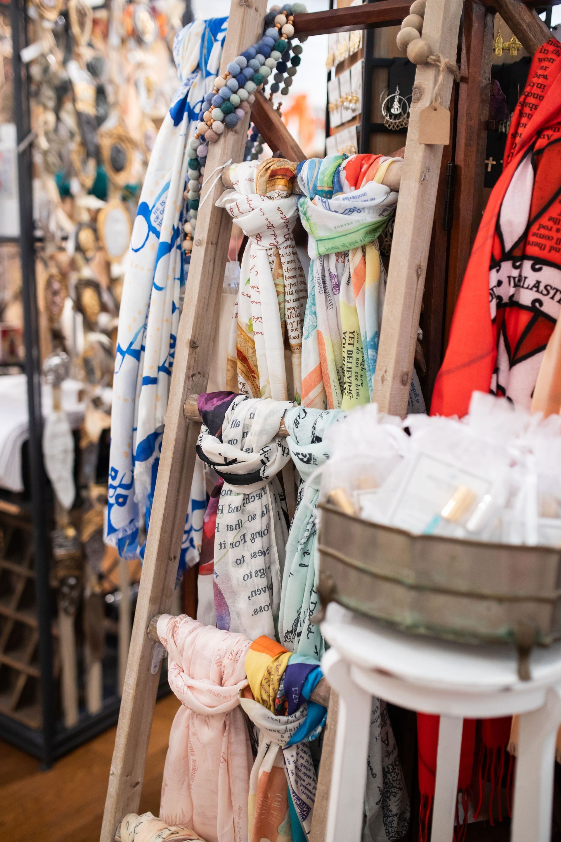 Scarves draped on a wooden ladder, displayed with a basket on a stool. Colorful and varied patterns.