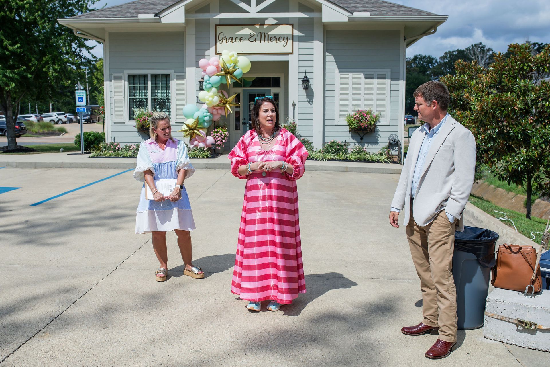 Three people stand outside a building with balloons. Woman in pink dress speaks, two look on.