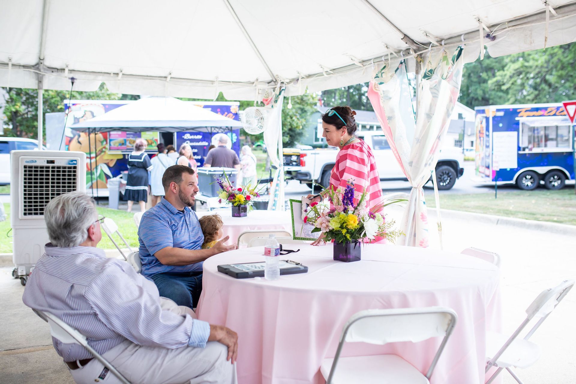 People gathered at tables under a tent at an outdoor event, with food trucks in the background.
