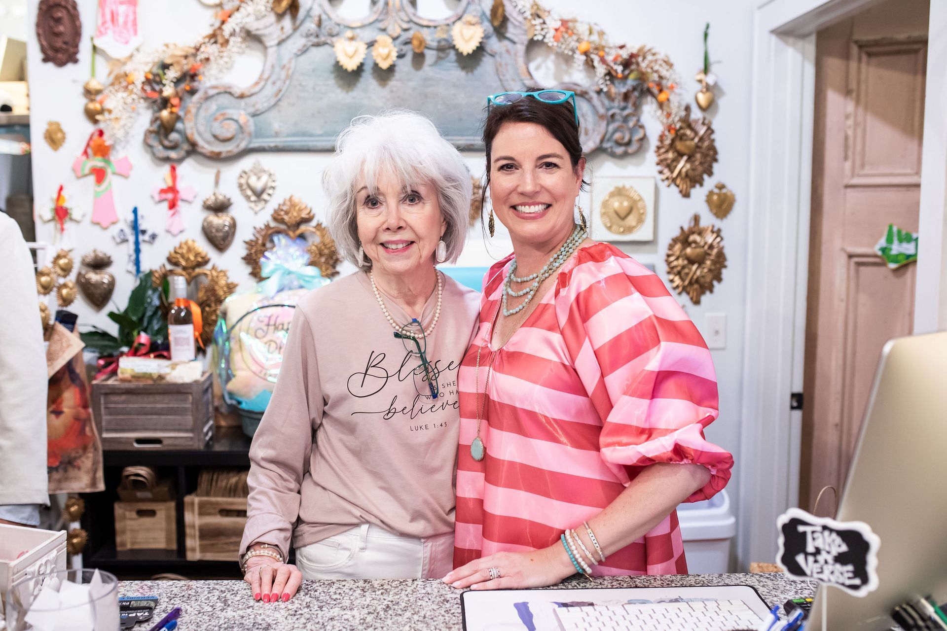 Two women smiling behind a store counter. One wears a striped top, the other a light-colored sweatshirt. Shop interior in background.