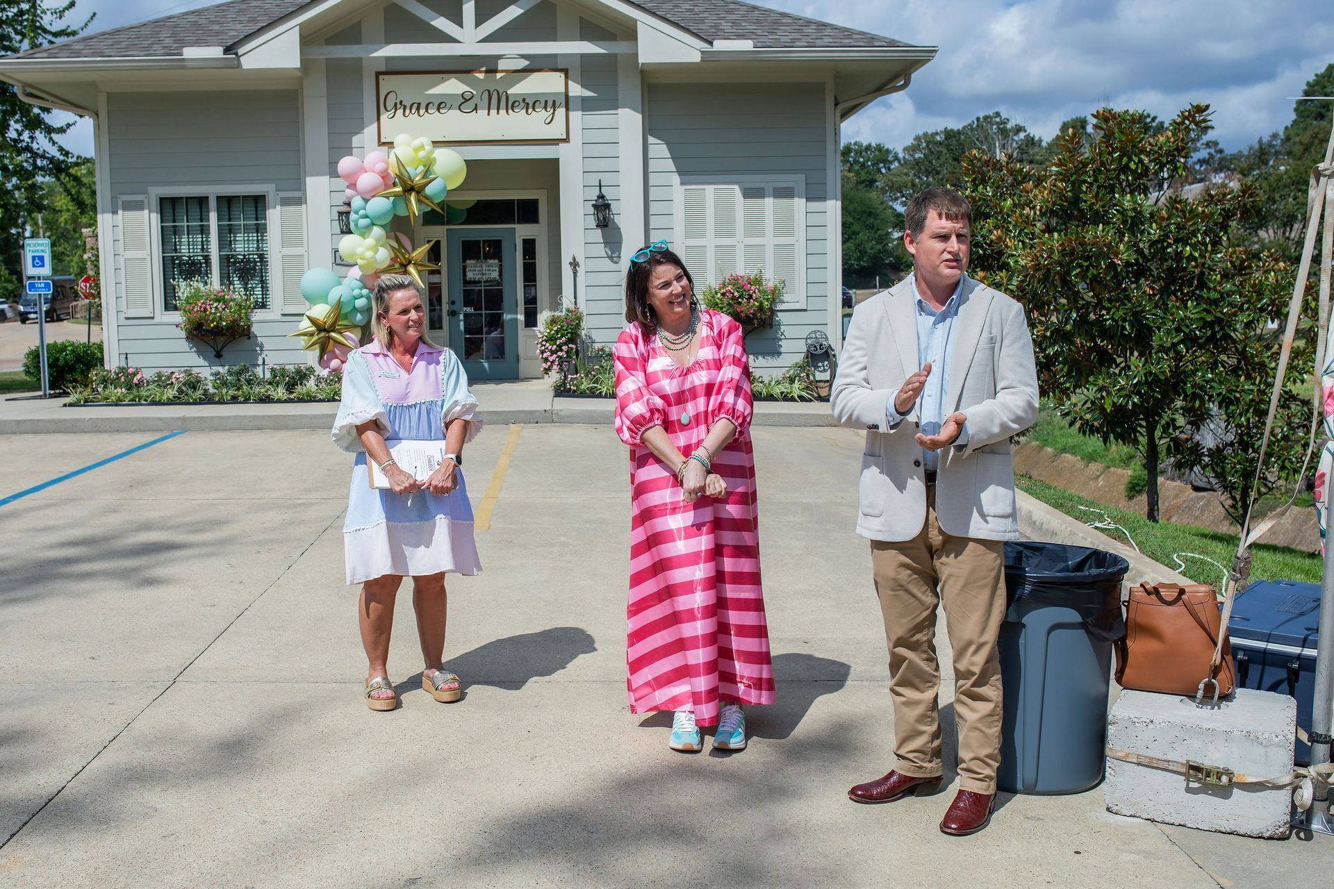 Three people standing outside a building with balloons. The woman in the center wears a pink dress. A man gestures.