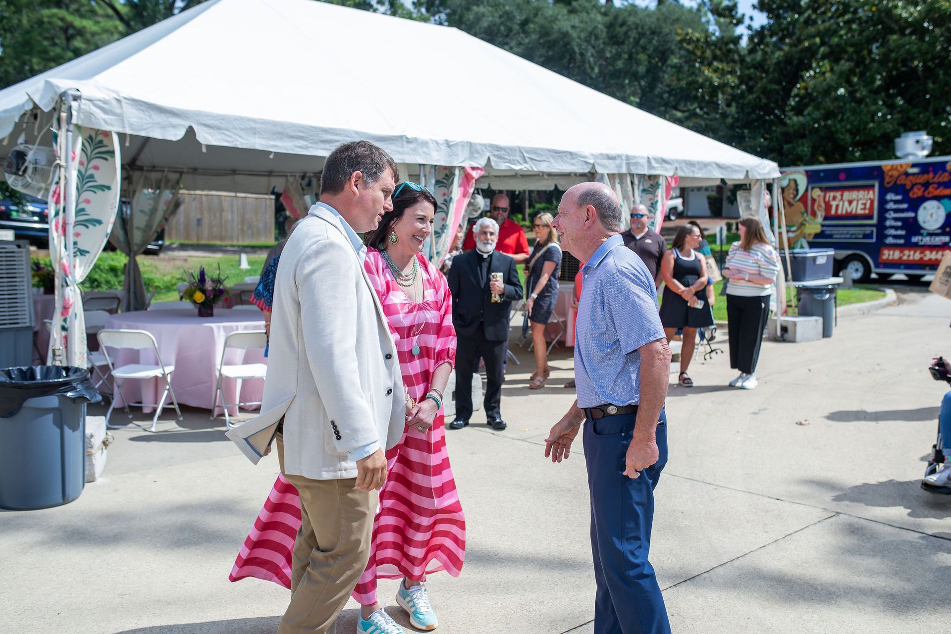 People converse under a white tent at an outdoor event. A food truck is visible.