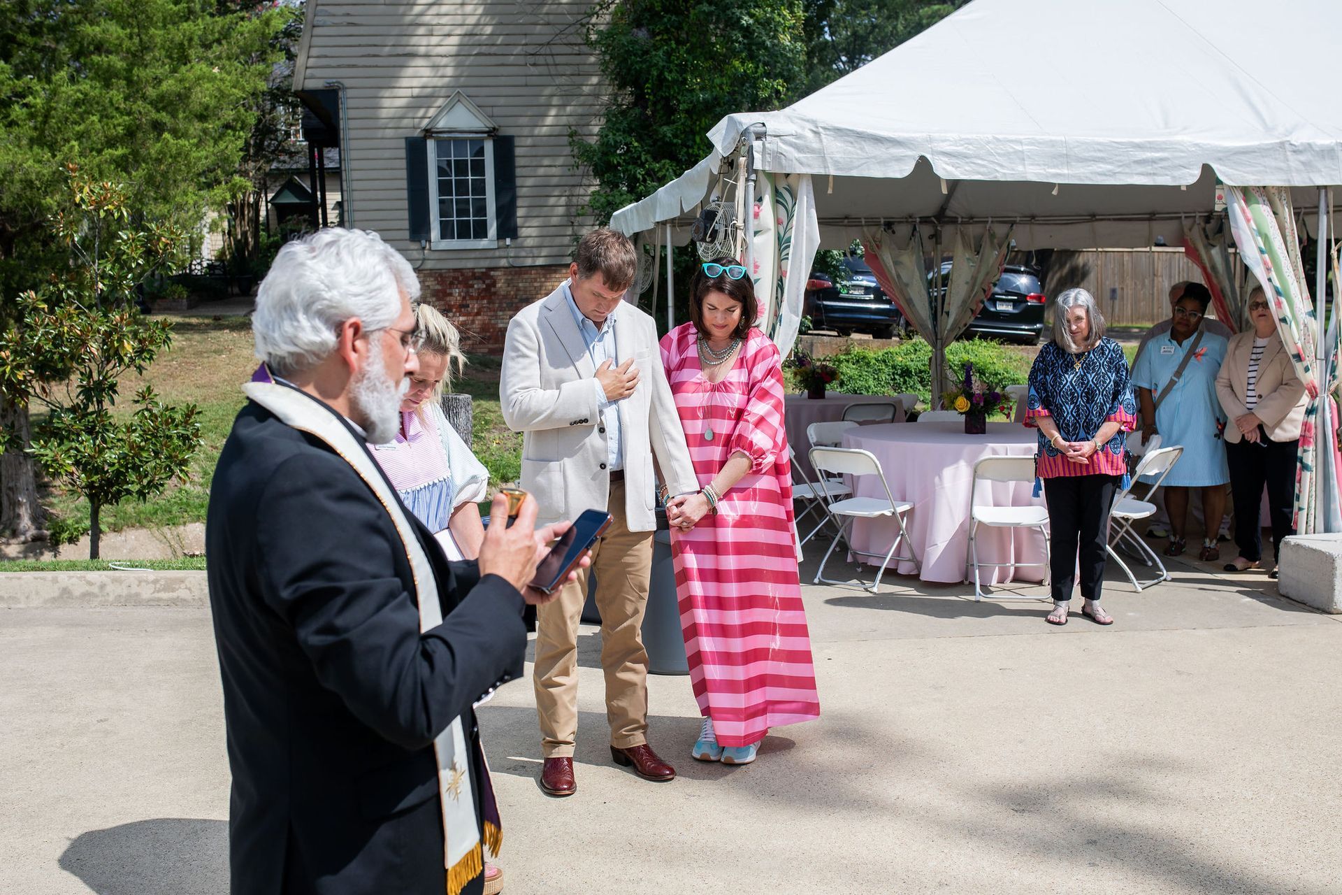 Wedding ceremony outside: couple with officiant under a tent, guests watching.