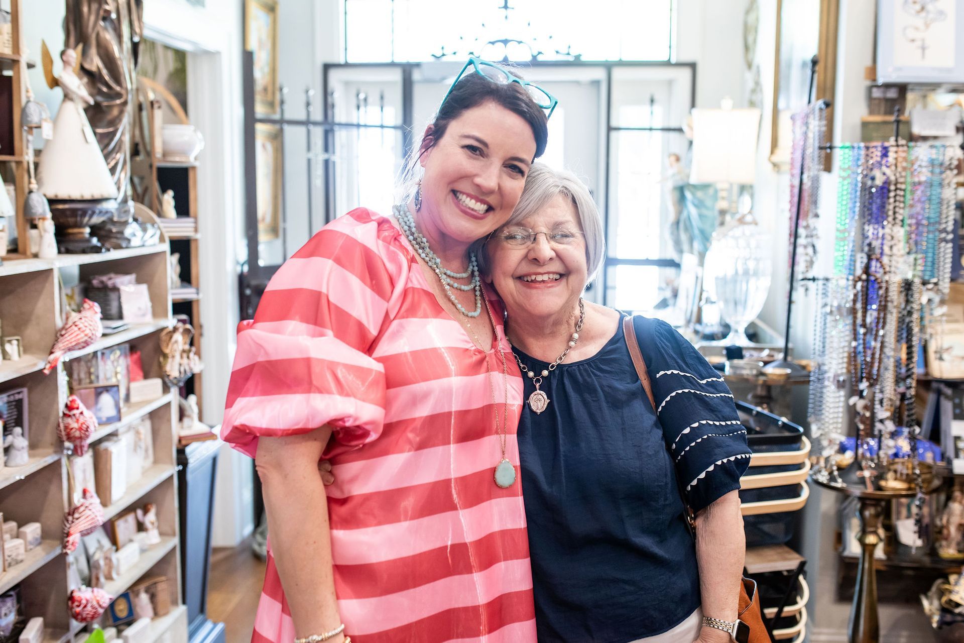 Two women smile, hugging in a well-lit shop, surrounded by merchandise. One in pink striped dress, the other in blue shirt.