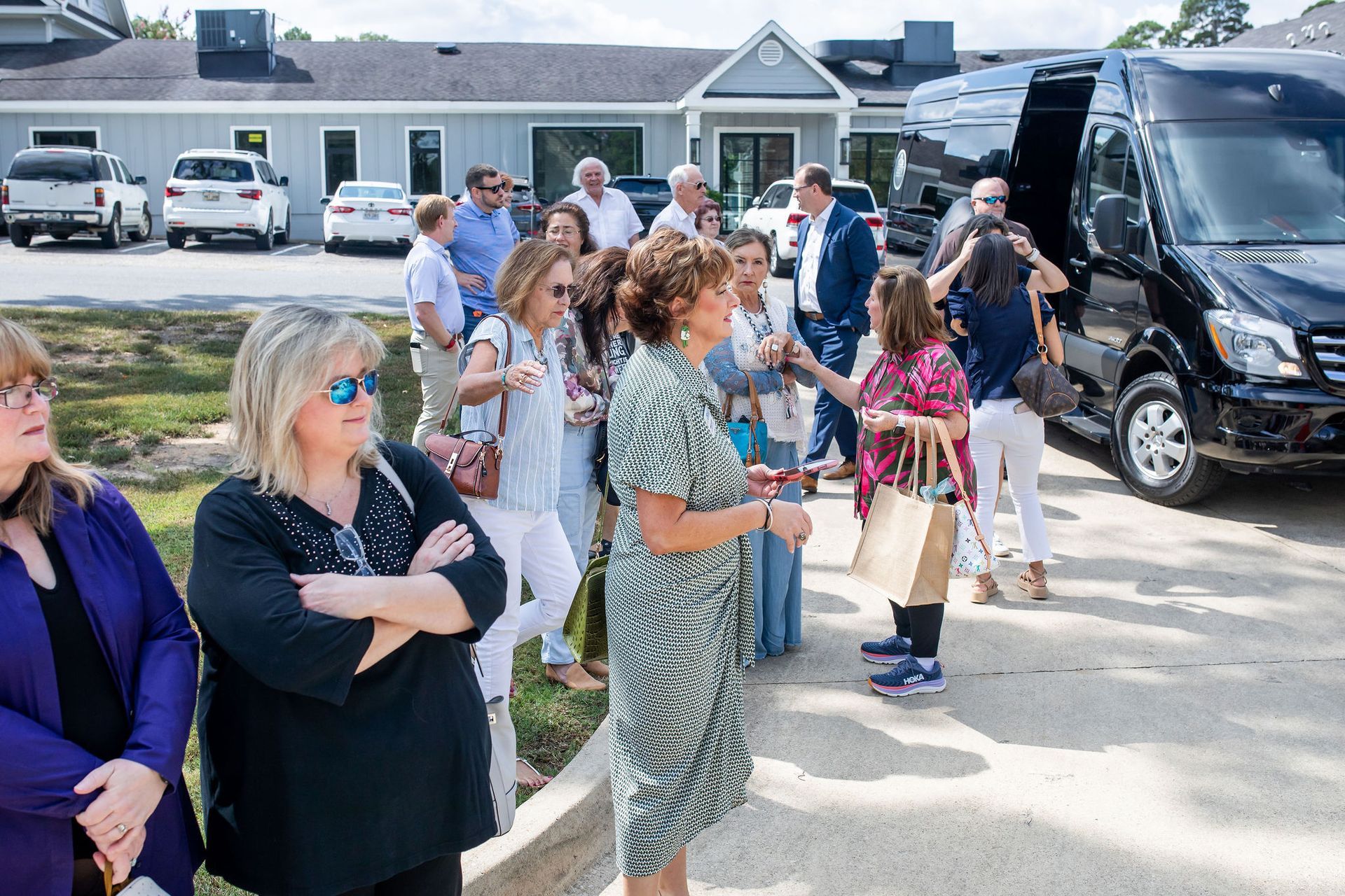 Group of people near a building and black van. Some are interacting, others are looking on a sunny day.