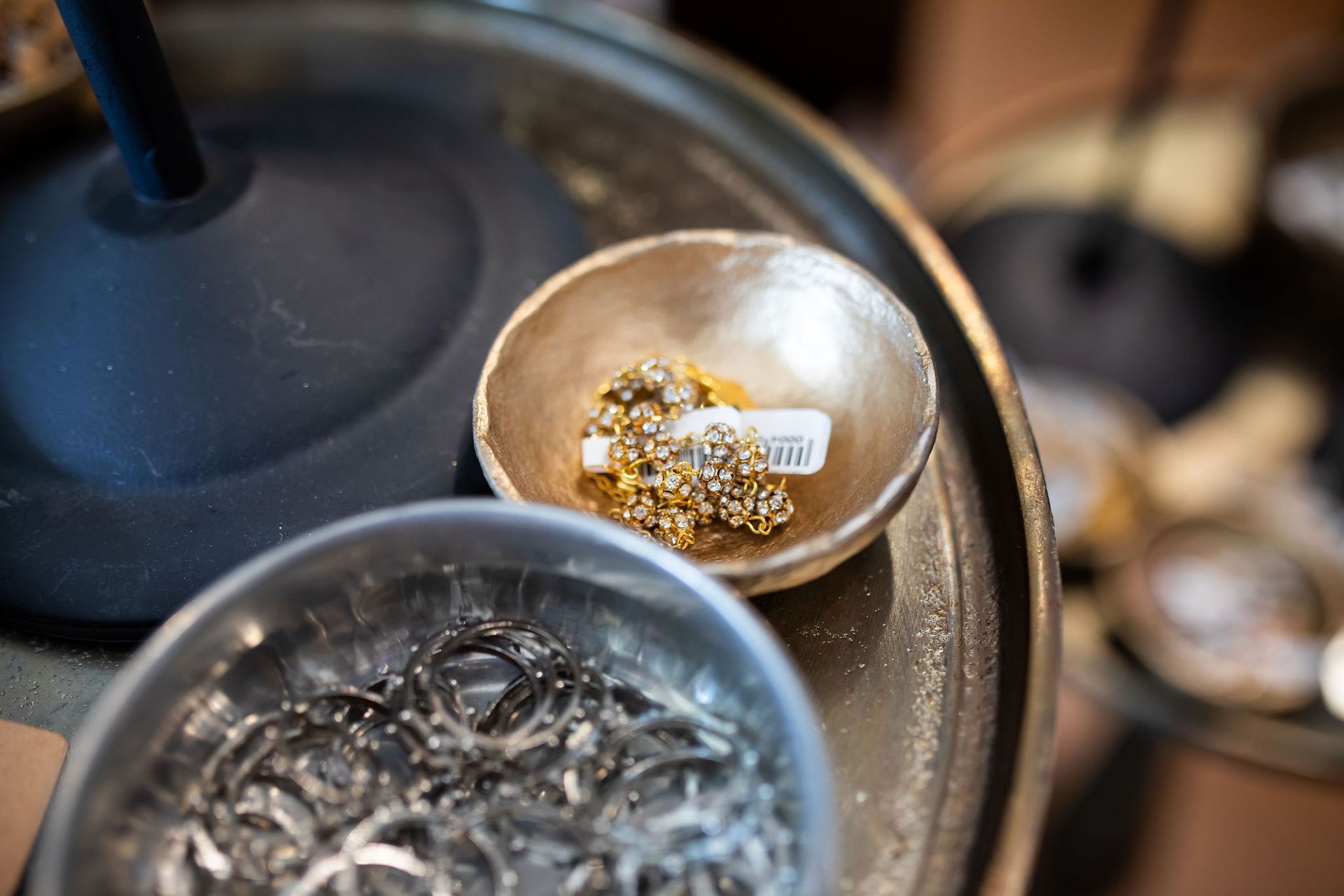 Small gold bowl with jewelry and price tag sits on a metal tray with another bowl of silver jewelry.