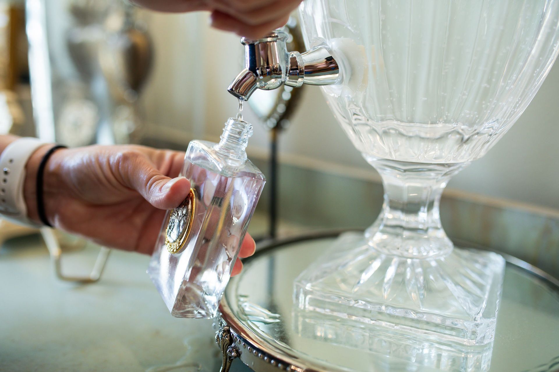 Hand filling a small glass bottle from a water dispenser on a mirrored tray.