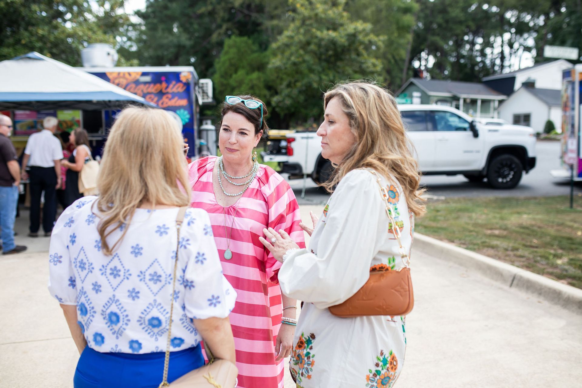 Three women chatting outdoors near food trucks and parked cars.