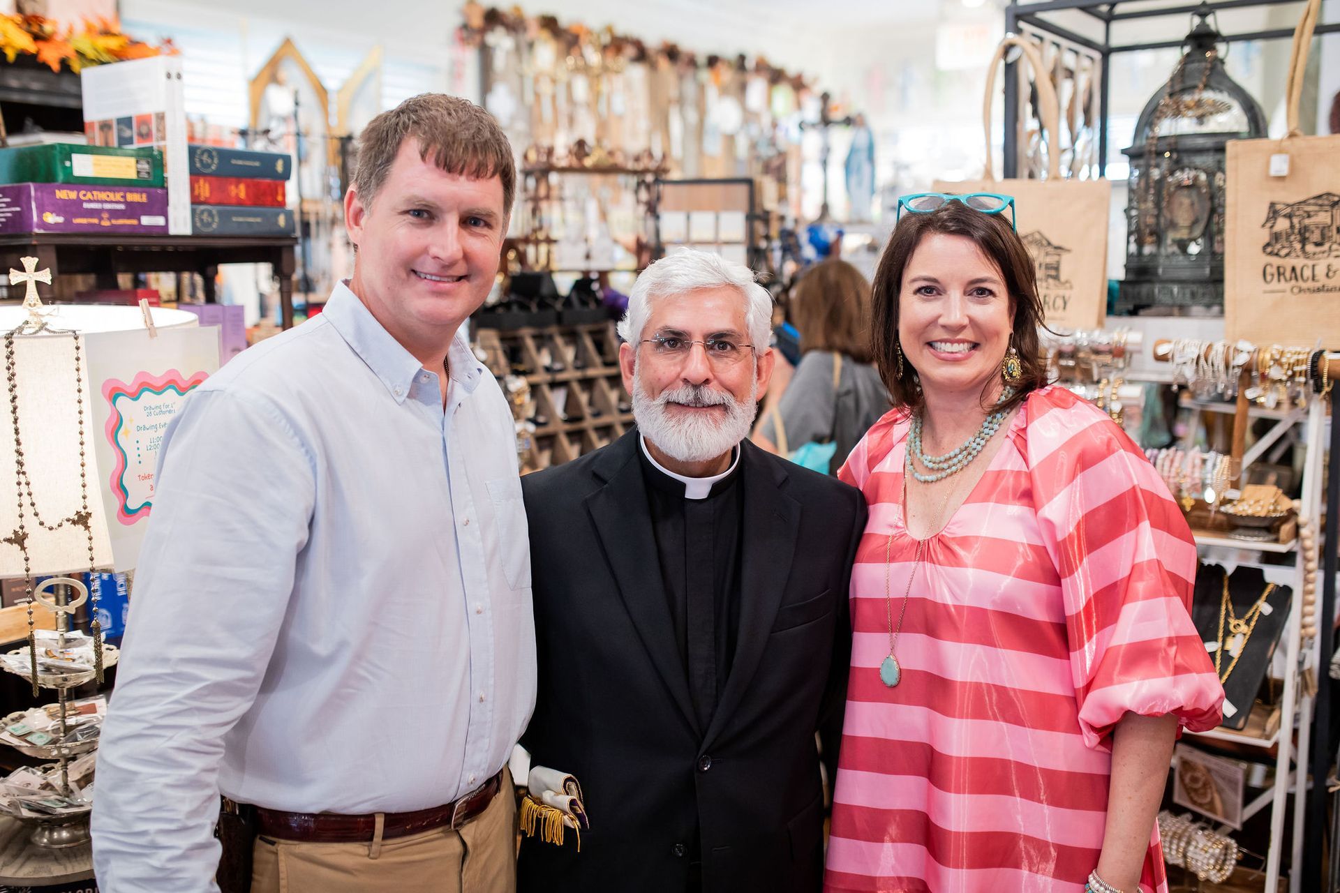 Two people flank a priest, smiling, inside a shop with various religious items.