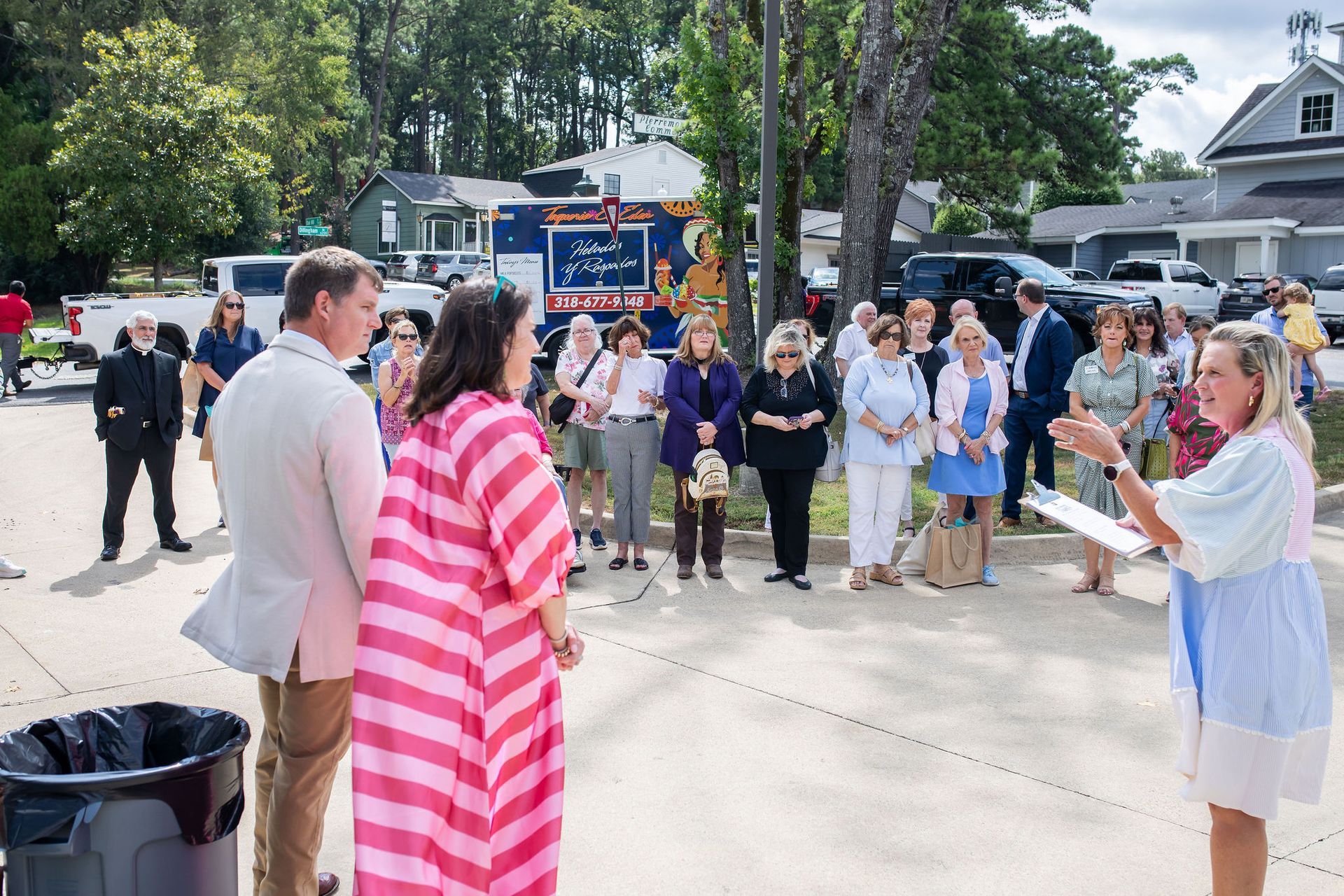Group of people gathered outdoors, some listening to a woman speaking, with a food truck in the background.