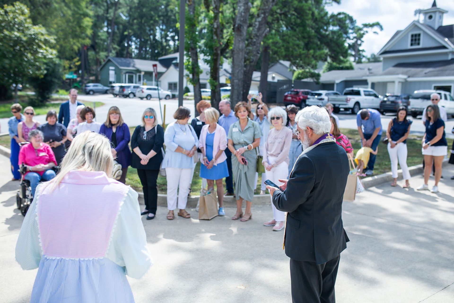 Group of people gathered outdoors, listening to a speaker. Building and trees in the background.
