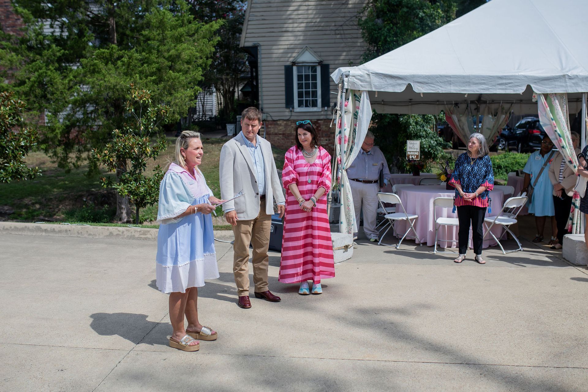 Group of people standing outdoors near a tent and a house. One person speaks, others listen.