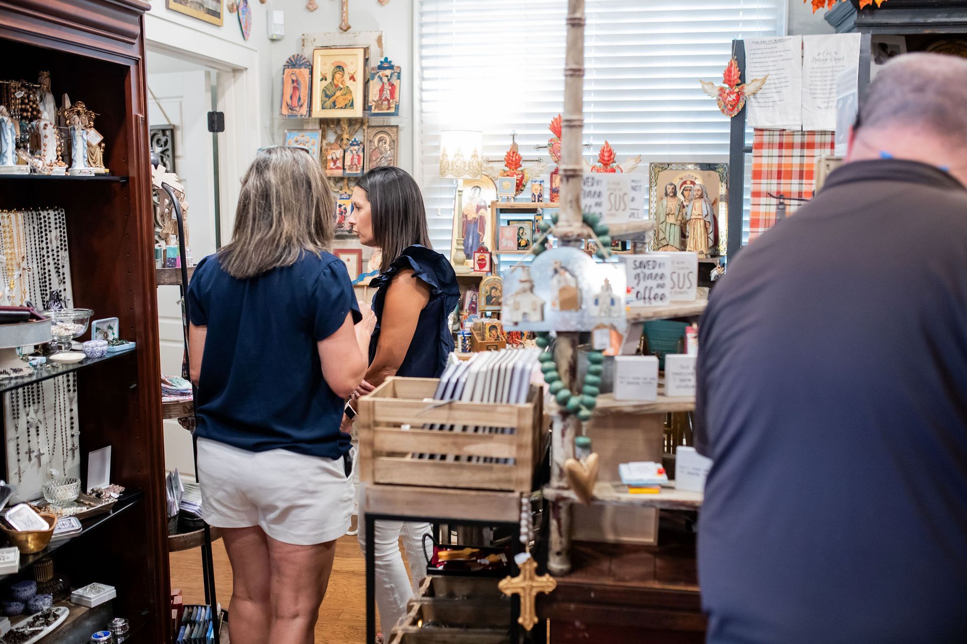 Two women browse items in a cluttered shop with religious artifacts; a man looks at the display.