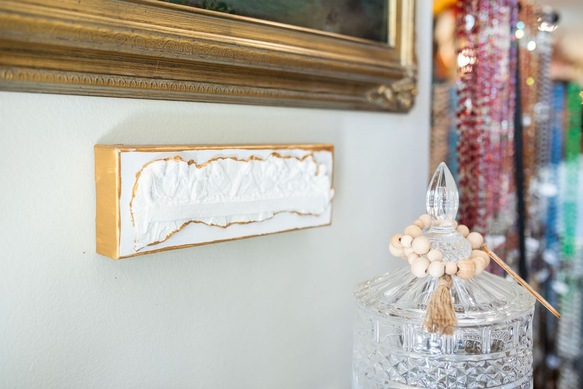 White textured art piece with gold frame on a wall, next to a crystal jar and a beaded item.