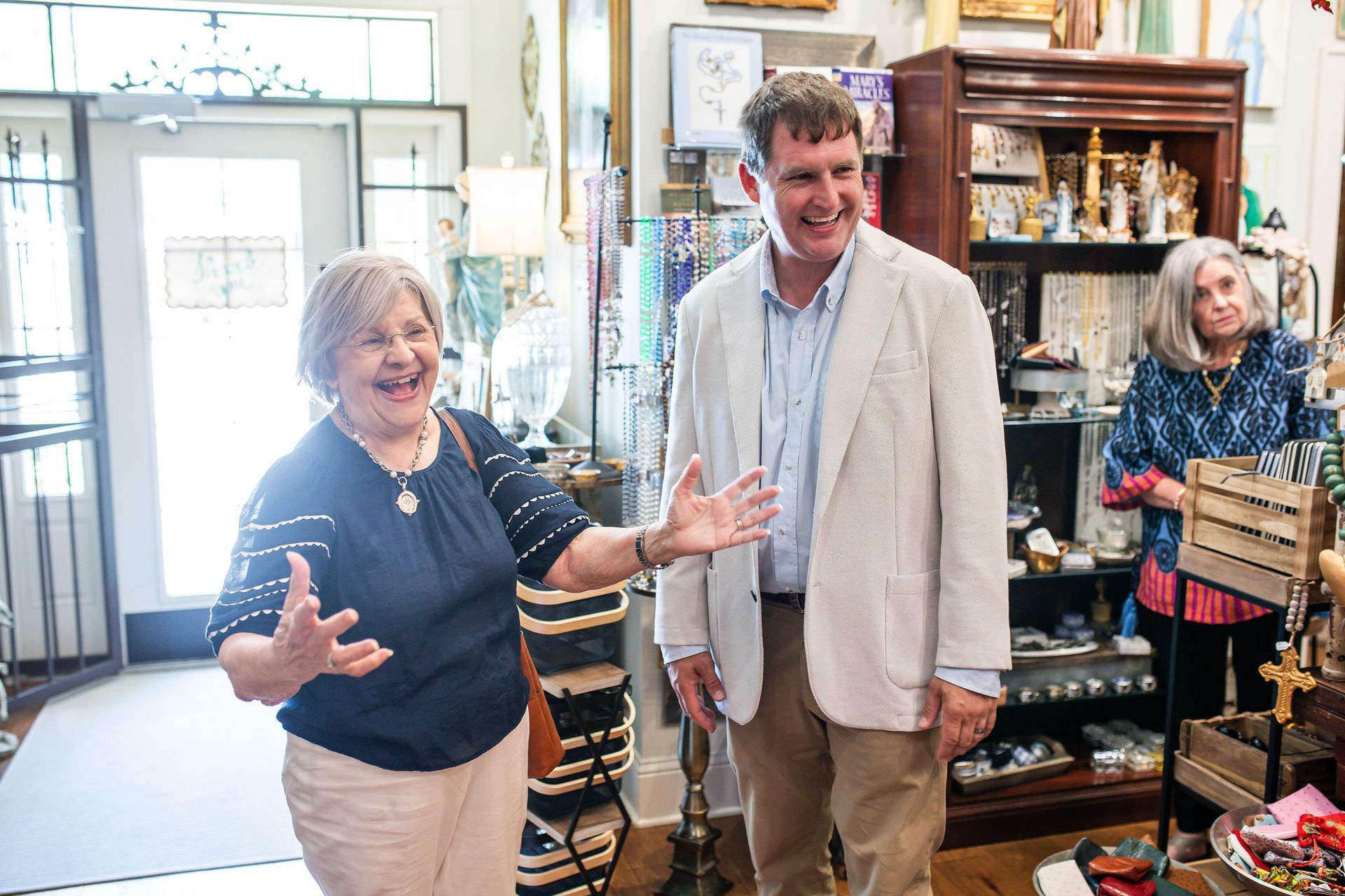 Woman gestures, smiling, to a man in a shop; a second woman looks on, shelves of items in the background.