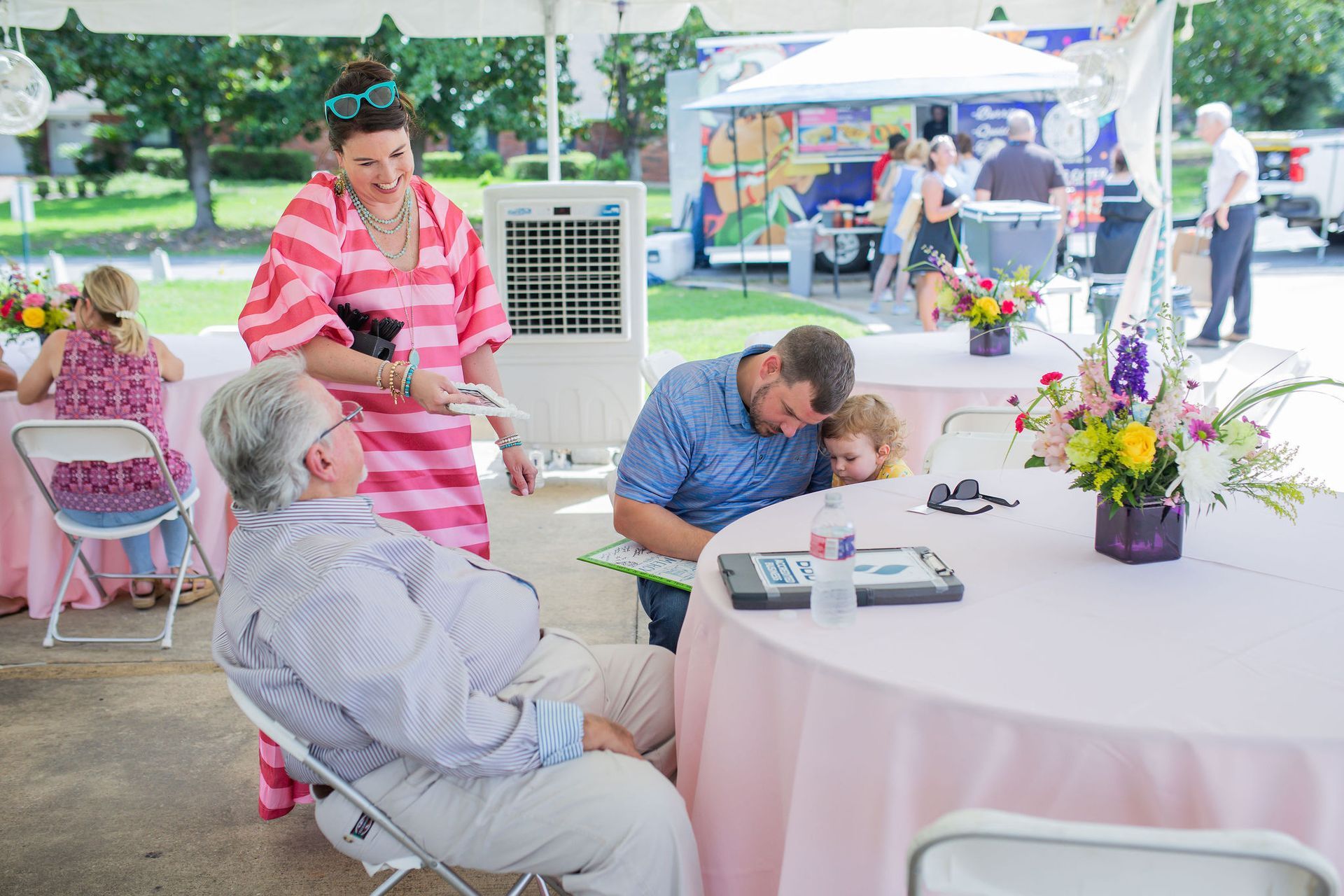 People at outdoor event under a tent, interacting around tables with pink tablecloths and floral centerpieces.
