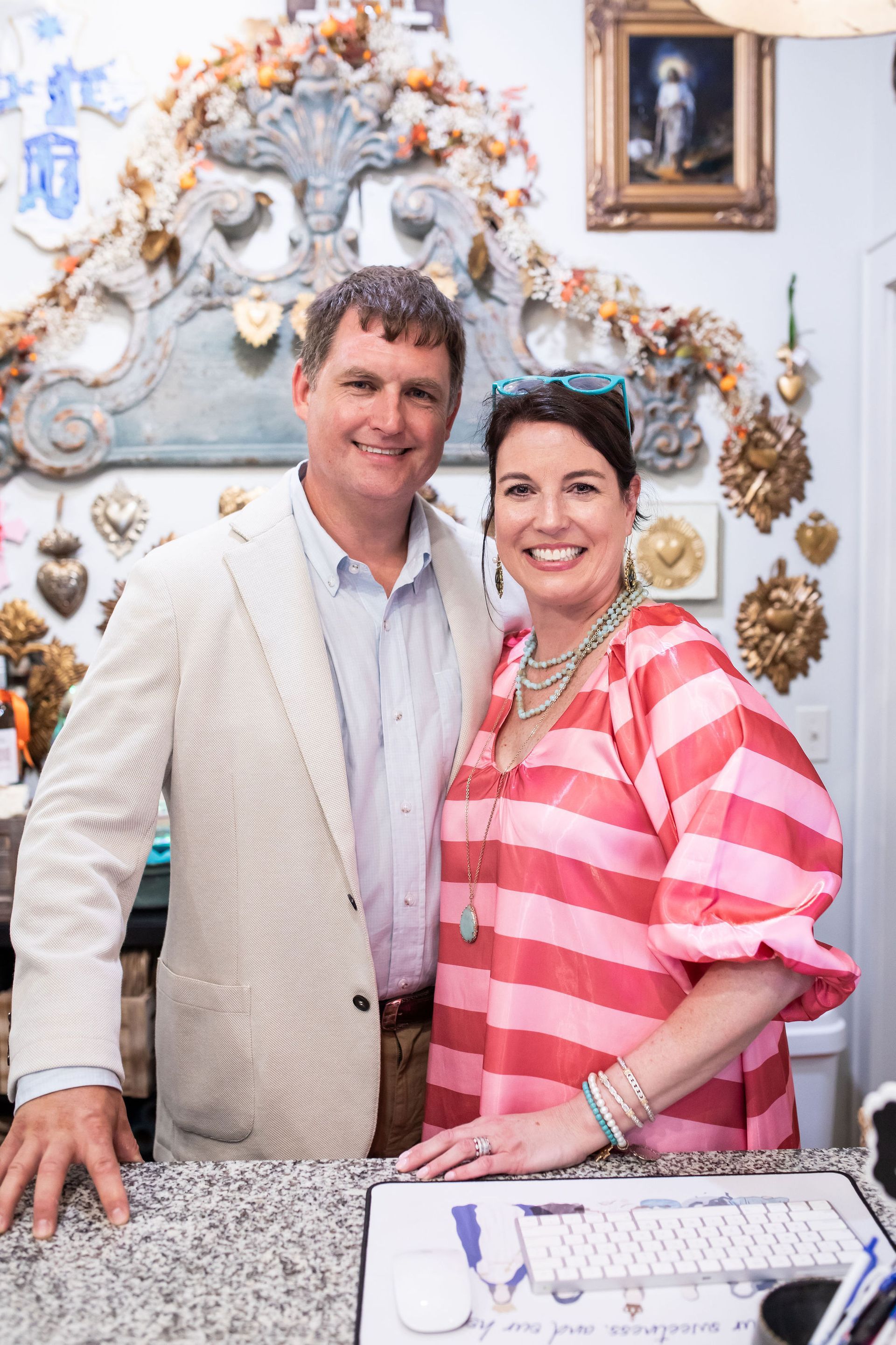 Couple smiling at the camera in a shop, woman in pink stripes, man in a tan jacket.