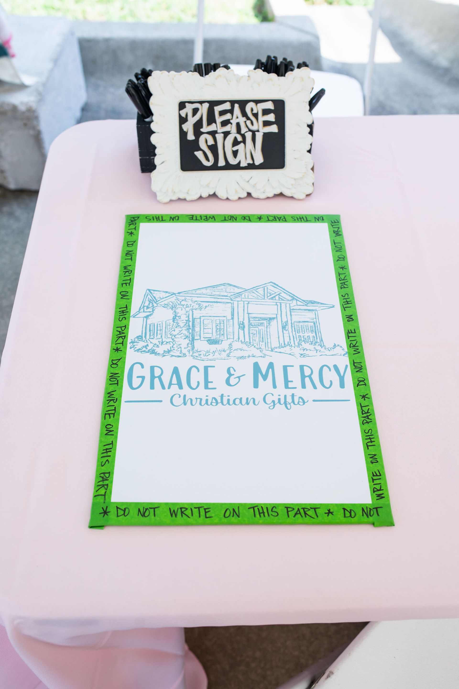 A sign-in table with a pink tablecloth and a “Please Sign” sign. 