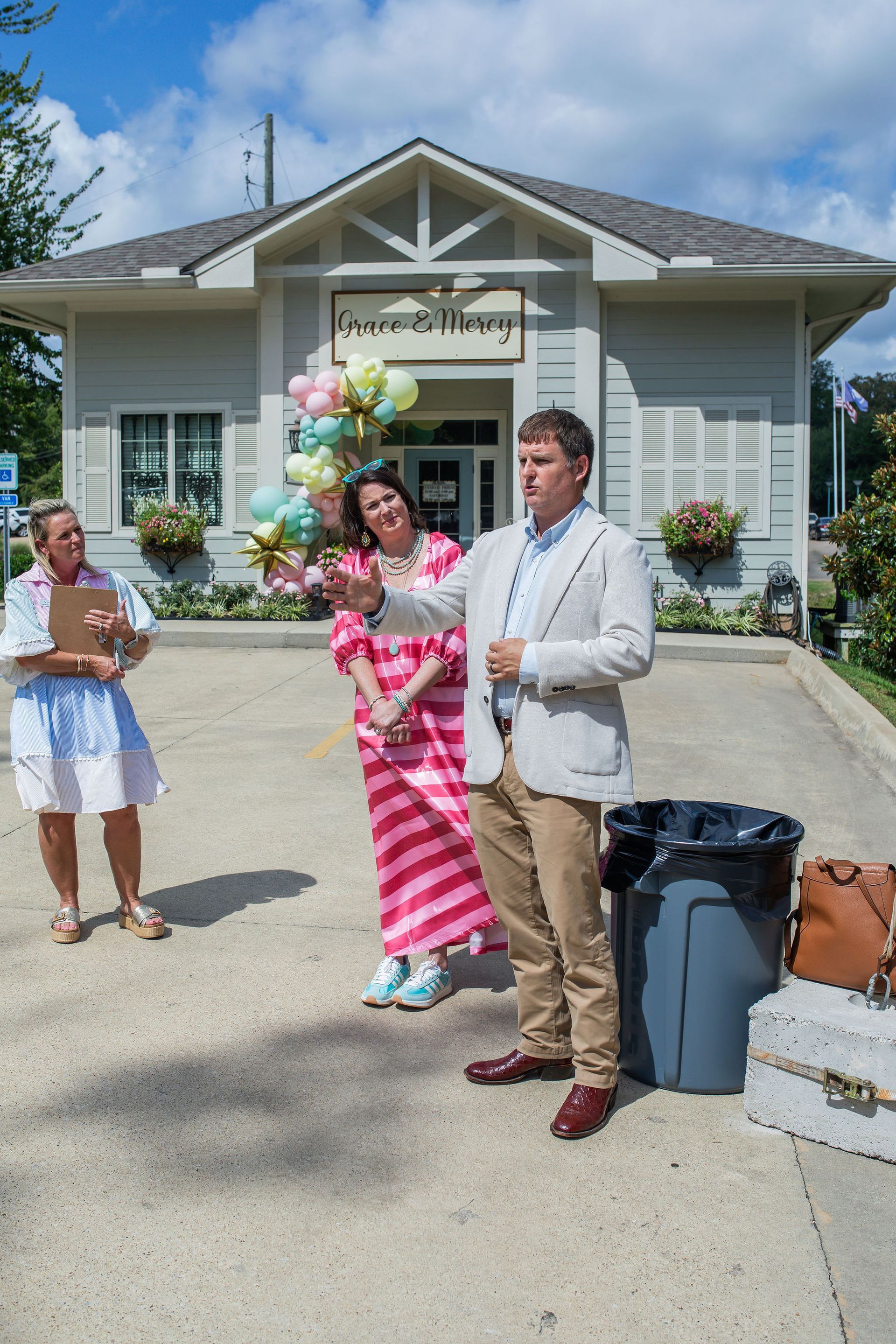 Three people outside a building. Man in blazer gestures, woman in stripes smiles, person with clipboard looks on. Building is light-colored.
