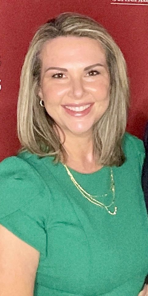 Woman in green dress smiles, wearing a gold necklace. Red background.
