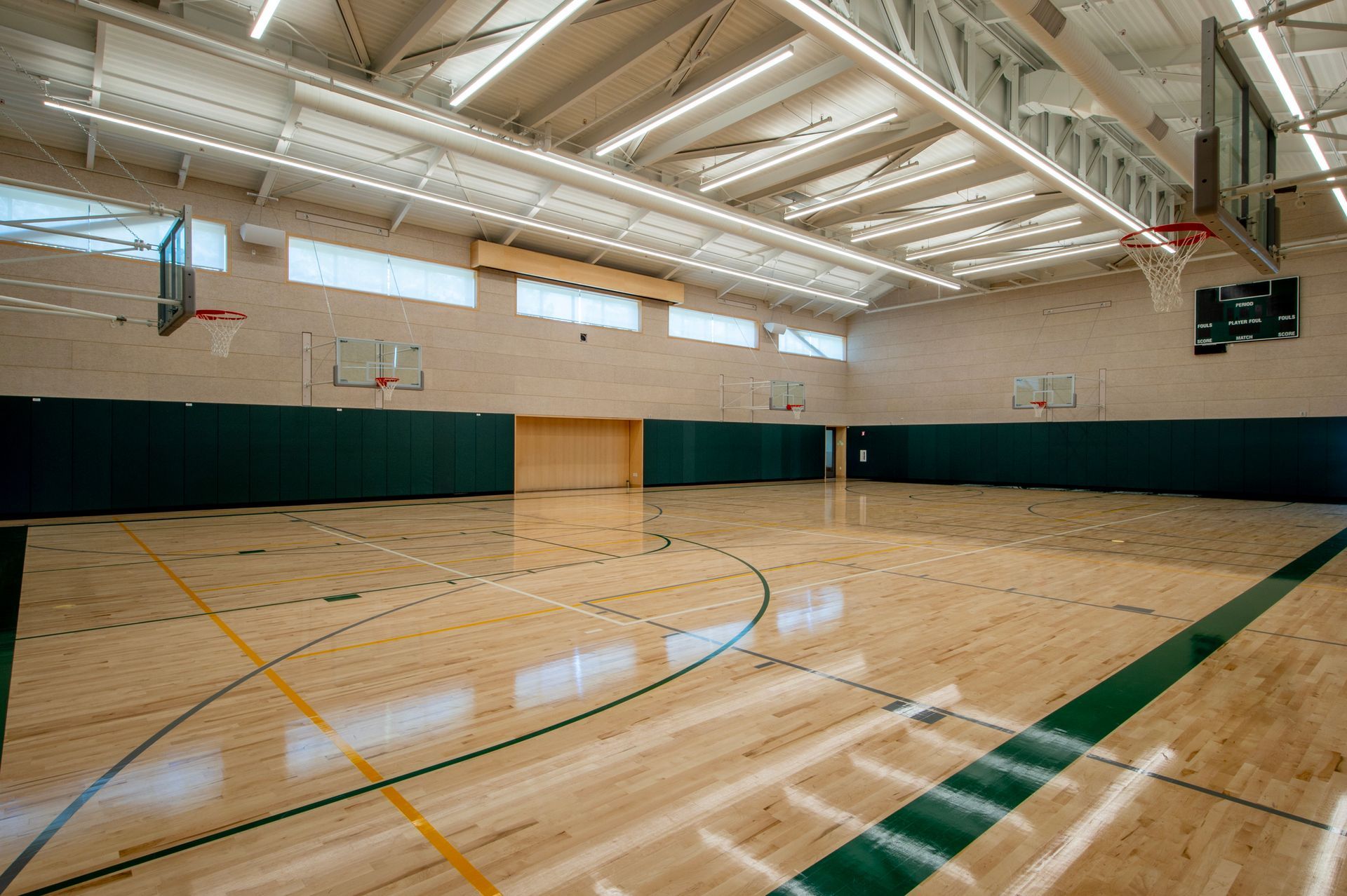 Empty indoor basketball court with wooden floor, green and white accents, and overhead lighting.
