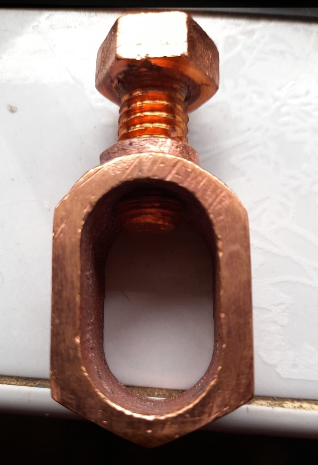 Tornillo de metal color cobre con un ojal hexagonal alargado sobre fondo blanco.