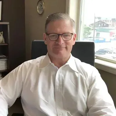 Man in a white shirt and glasses sitting in an office beside a sunlit window