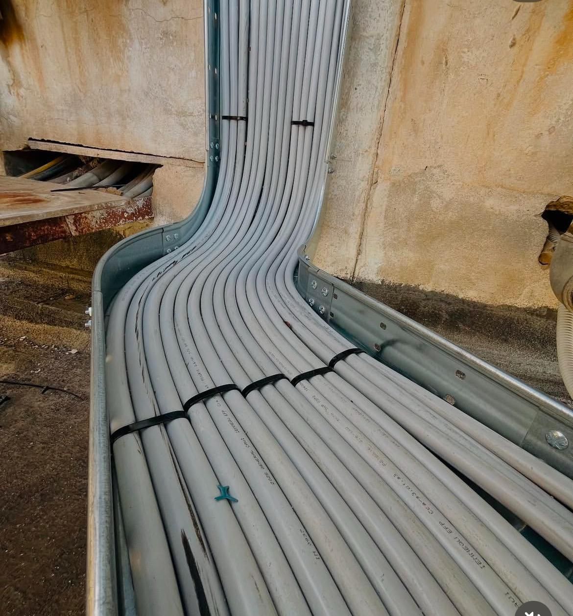 Cavi elettrici che sporgono dai fori nel muro; alcuni sono raggruppati, altri sparsi sul pavimento di cemento.