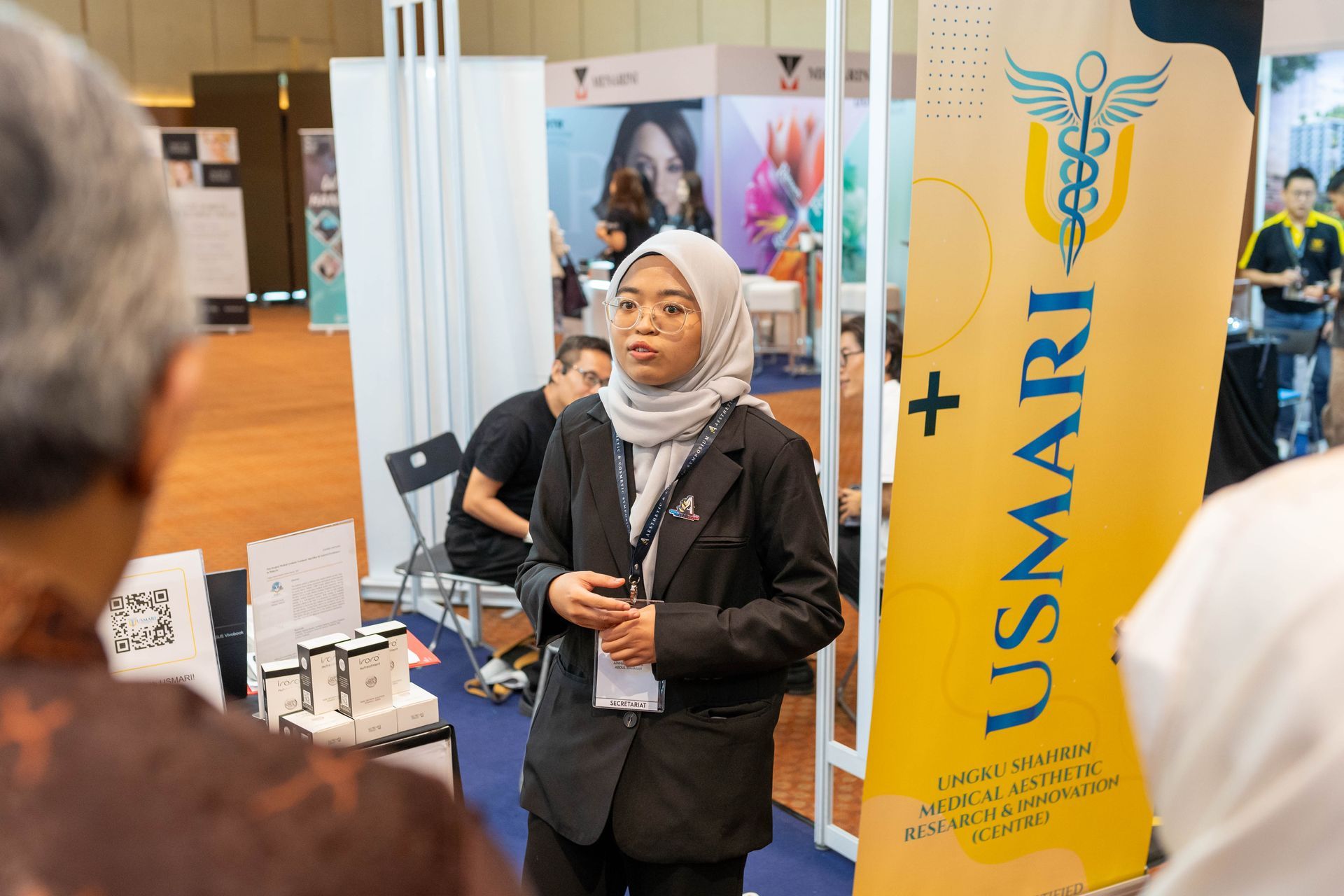 Woman in hijab speaking at a booth with USMARI signage, at a fair.
