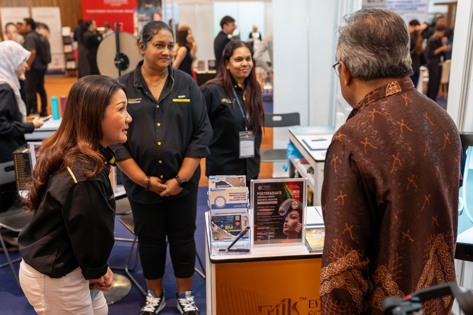 People at a booth in a convention hall; two women and a man talking, another woman looks on.
