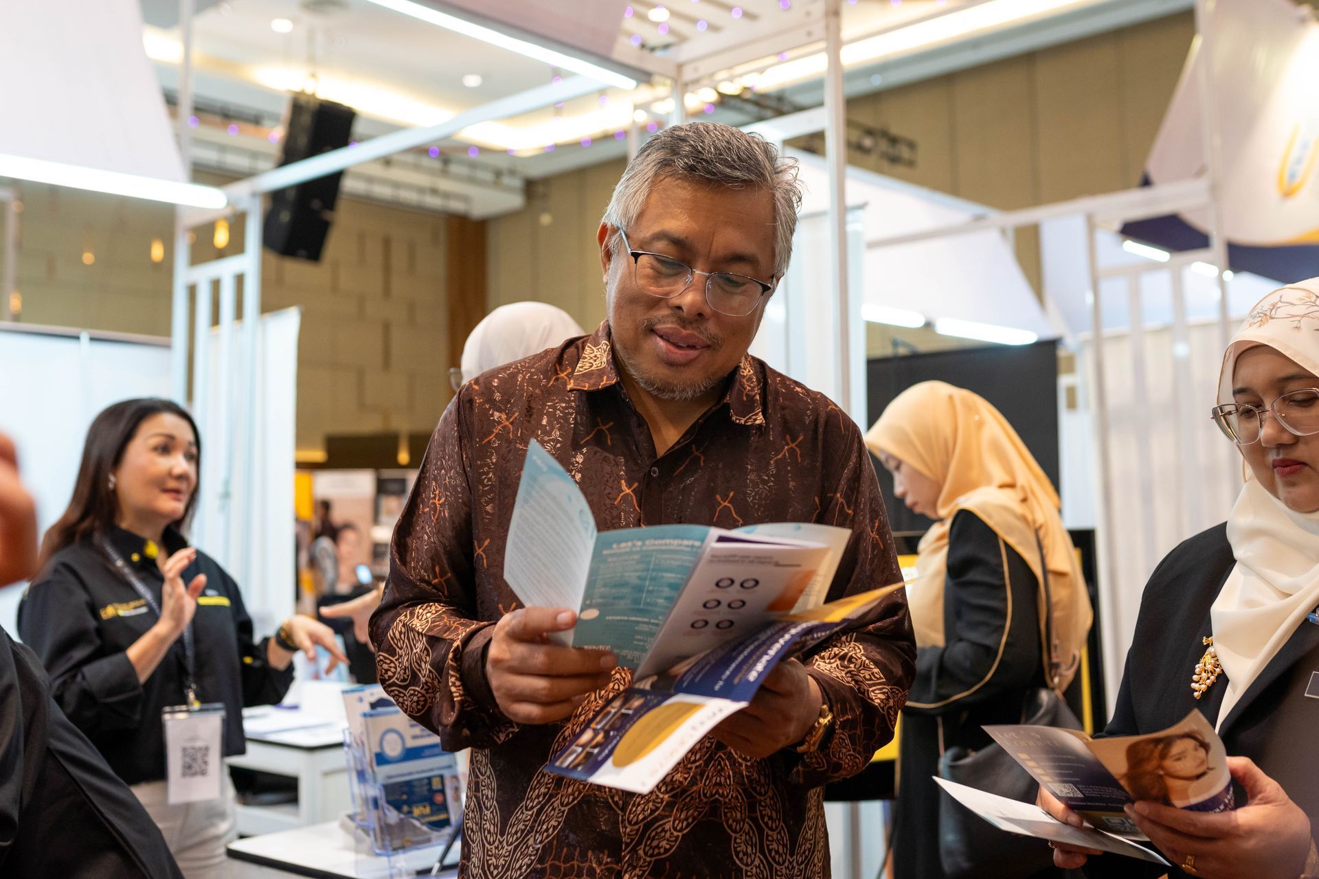 Man in patterned shirt reads brochure at an event. Other attendees observe nearby.