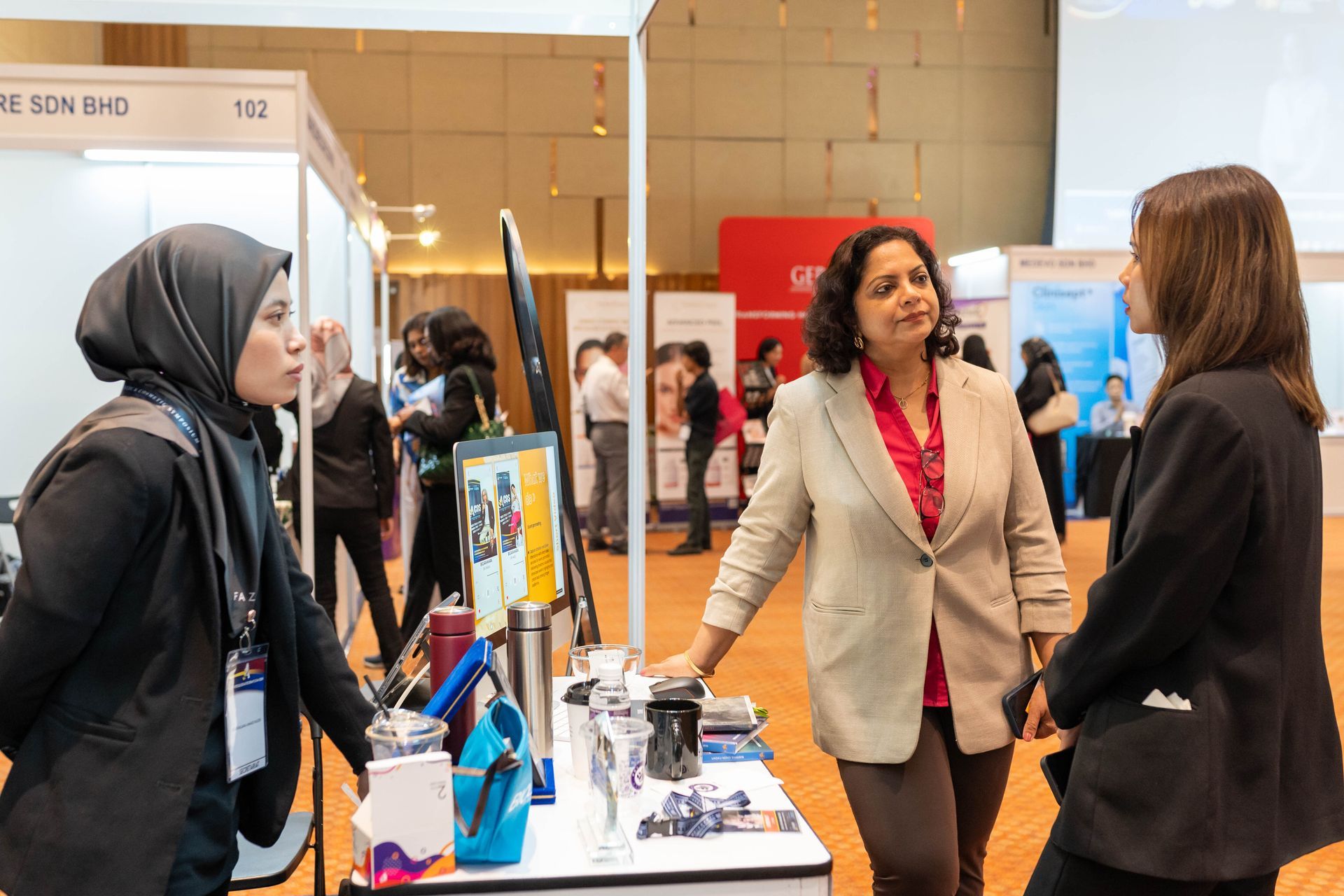 Three women converse at a trade show booth; a woman in hijab, one in a blazer, and another facing away.