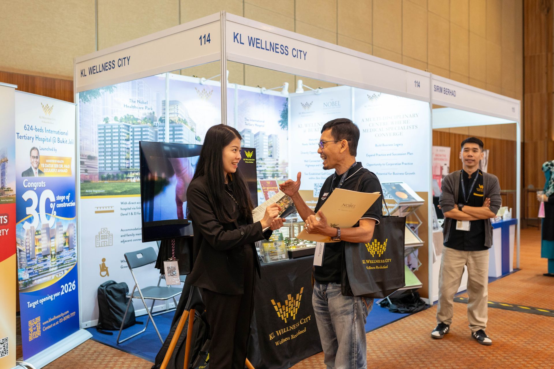 Two people shaking hands at a trade show booth, other people and displays are visible in the background.