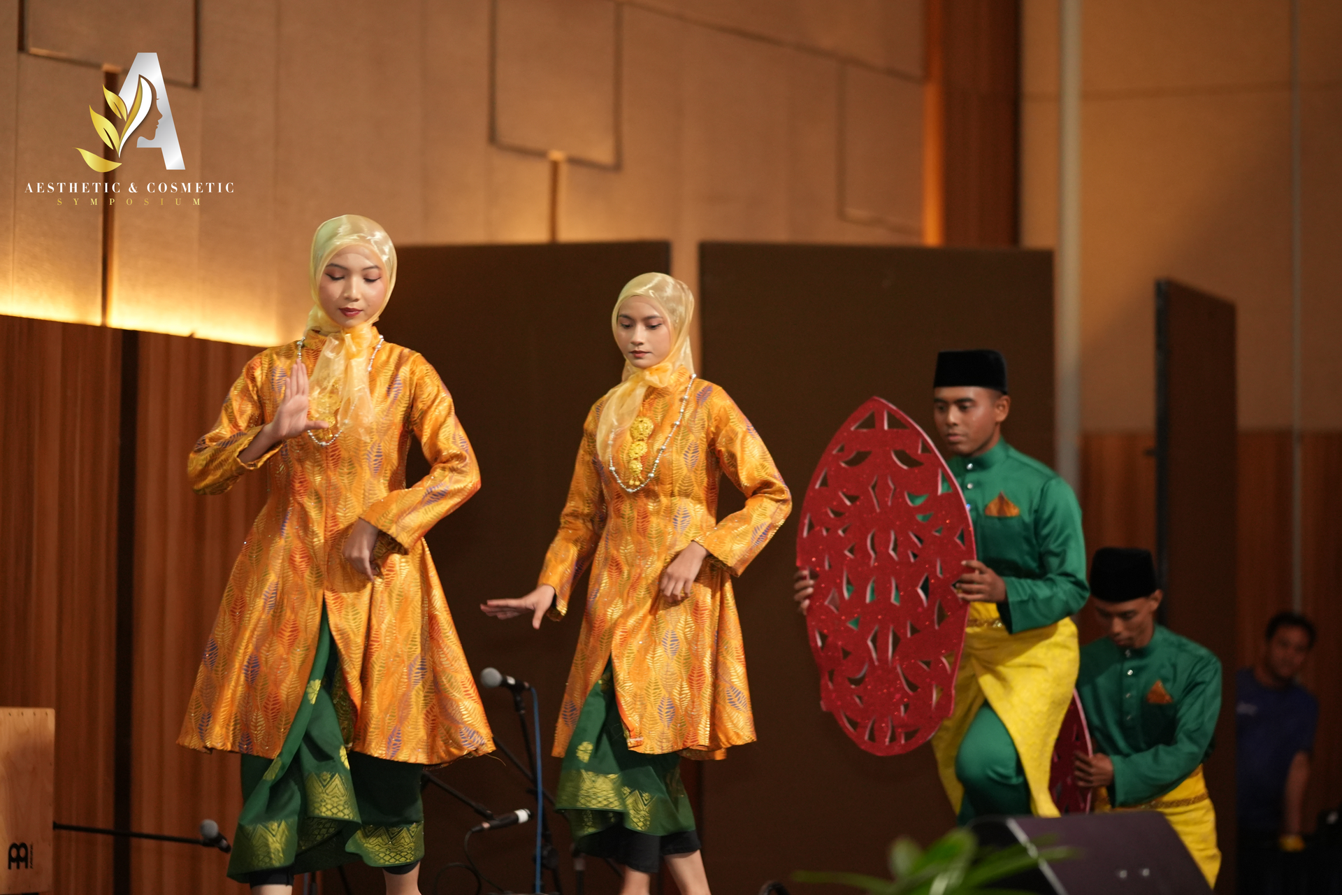 Two dancers in gold jackets and green skirts perform onstage with two musicians holding a decorative shield.