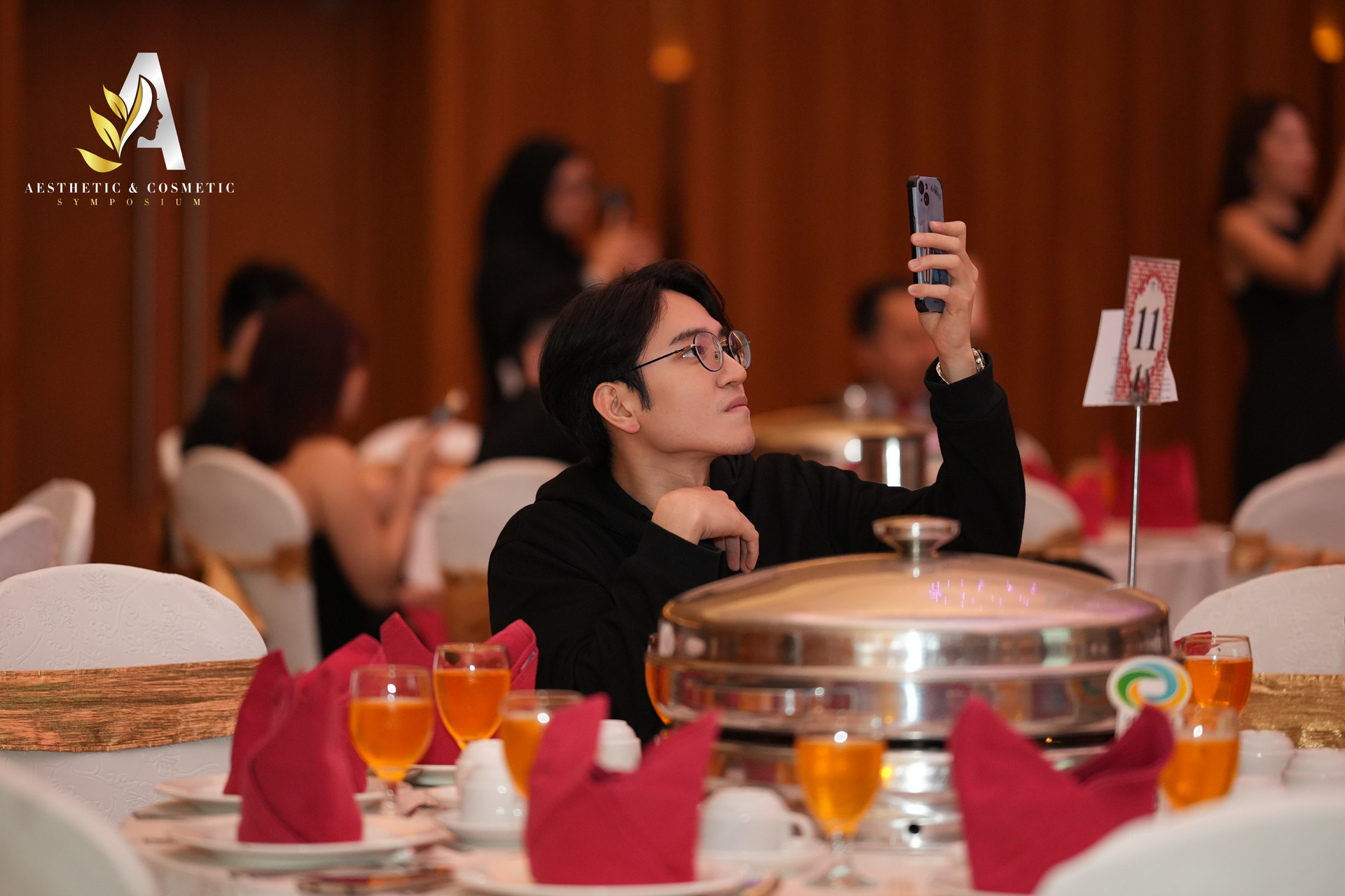 Man taking a selfie at a banquet table; formal setting with others, food, and drinks.