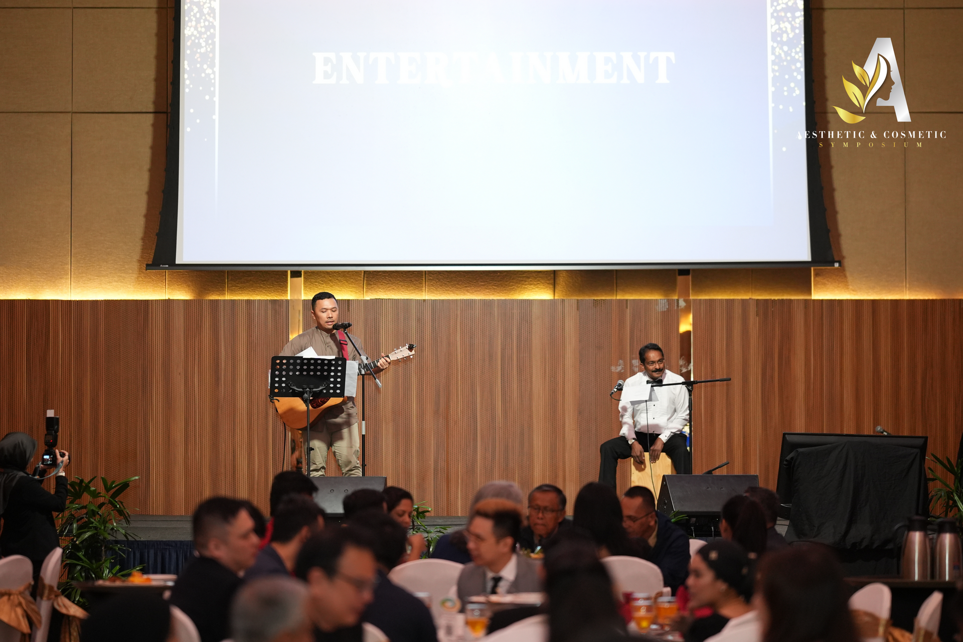 Two musicians performing on stage at an event. A screen displays 