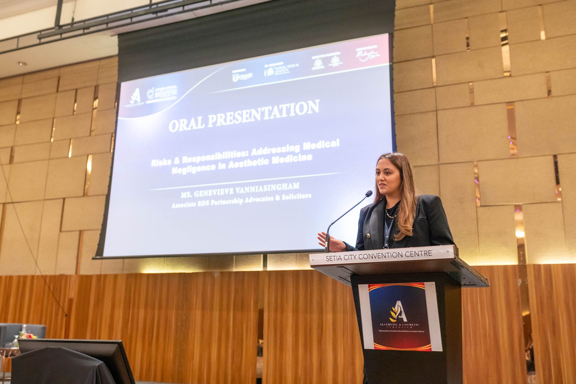 A woman gives an oral presentation on a stage. Presentation title displayed on screen.