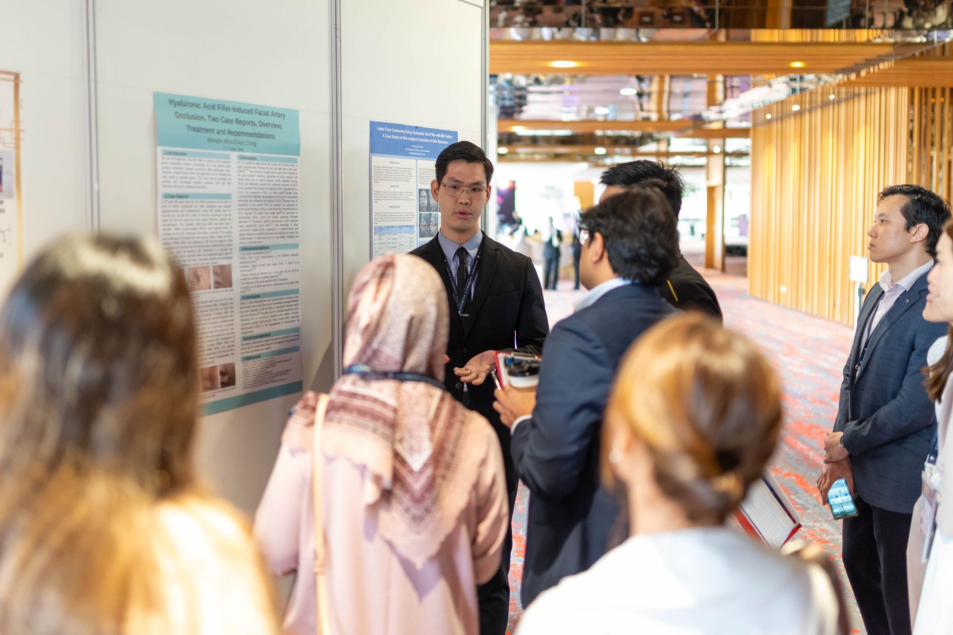People at a conference listening to a presentation next to a poster board. Indoors, natural light.