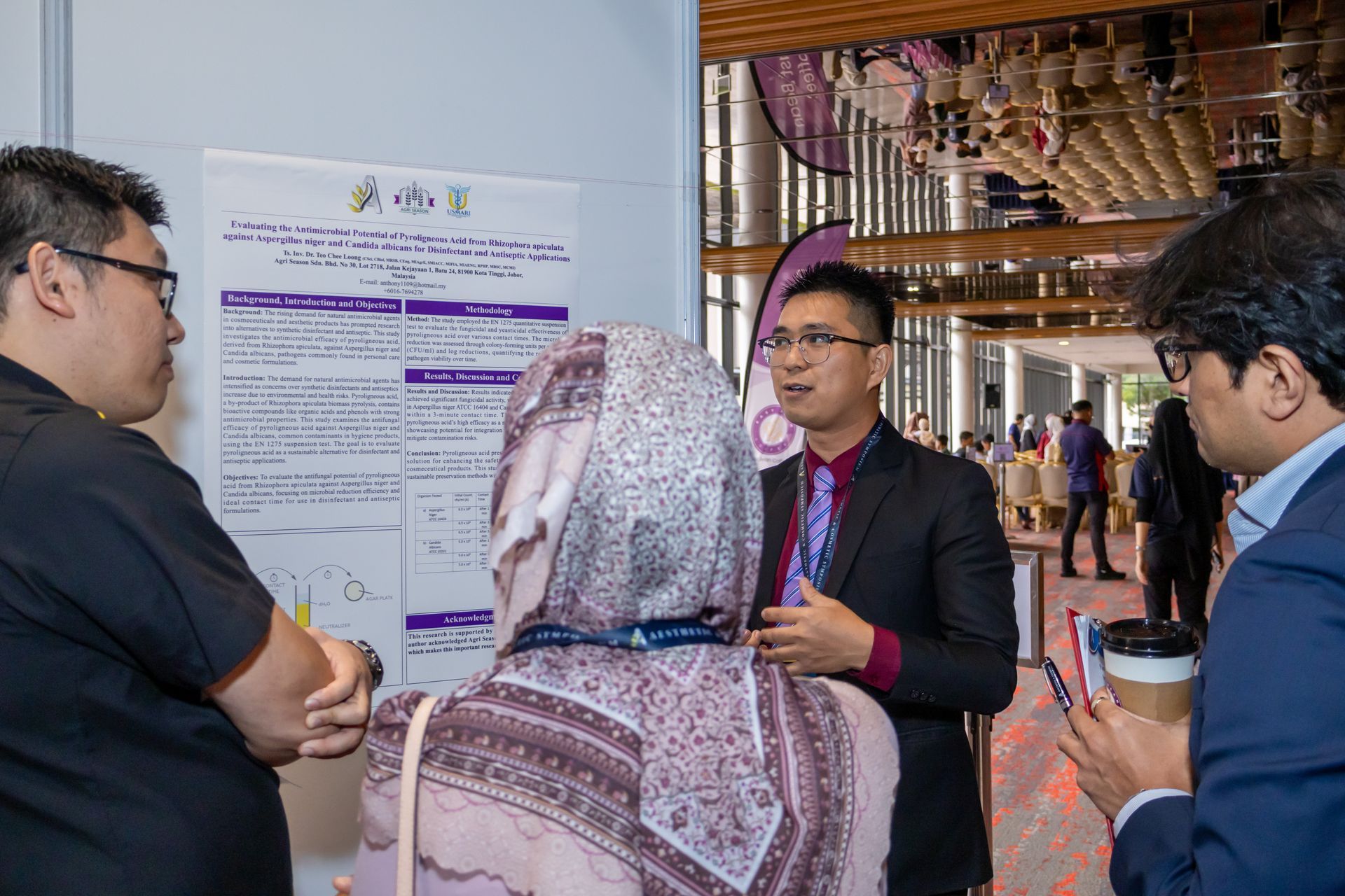 People at a conference presentation. Man in suit explains poster to attendees, one wearing a headscarf.