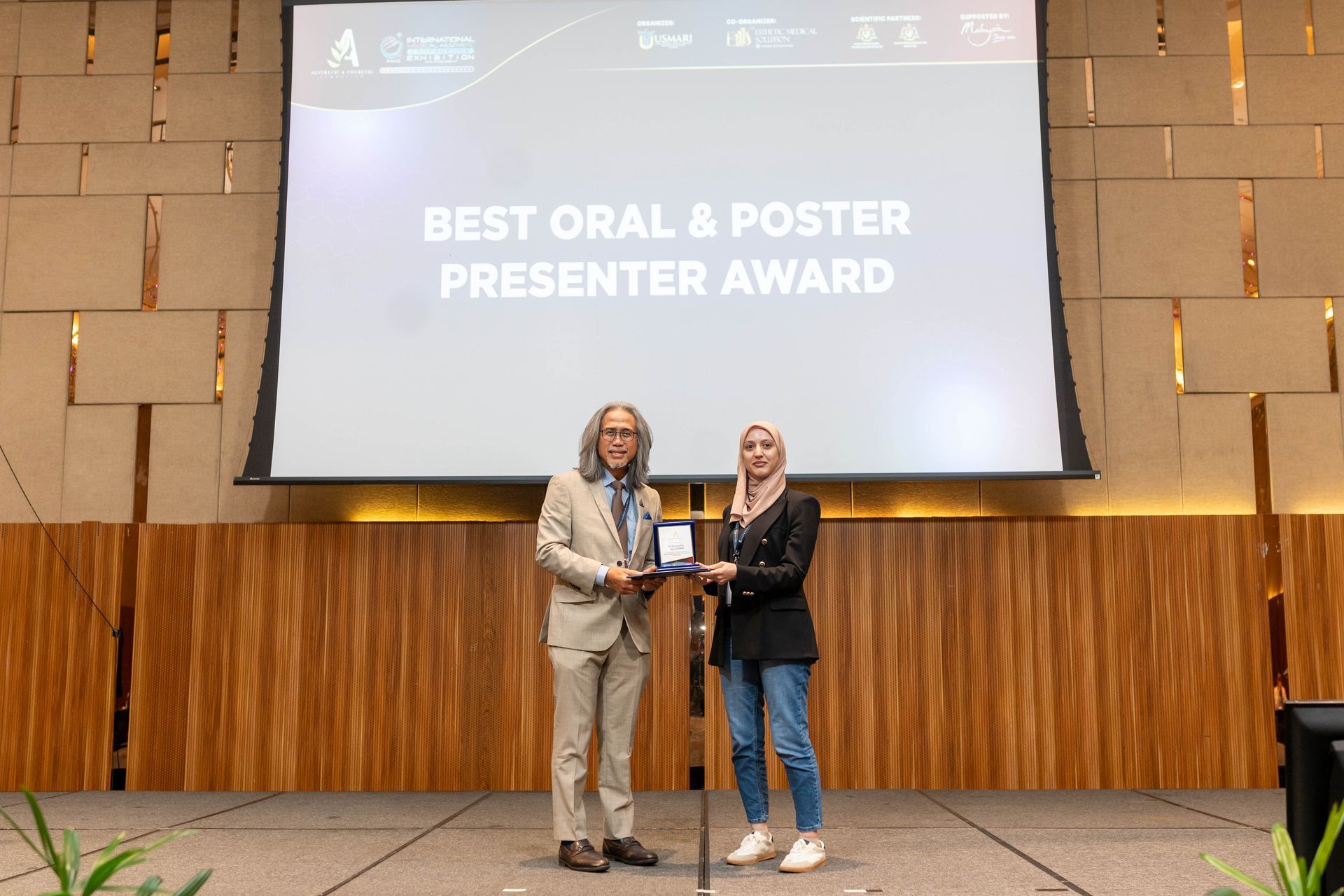 Man in suit presents award to woman in blazer on stage with backdrop.