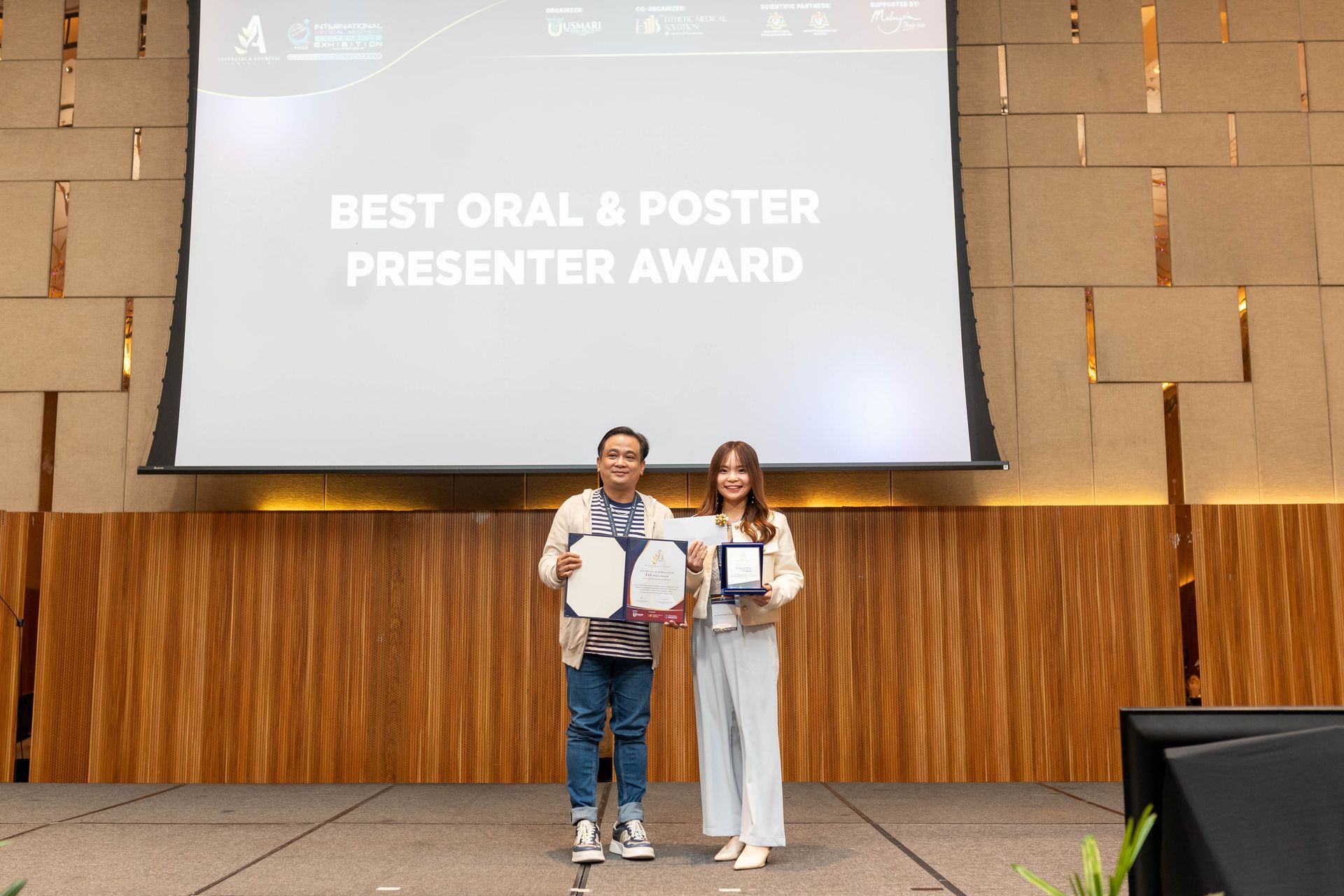 Two people holding awards on a stage. Text on screen: 