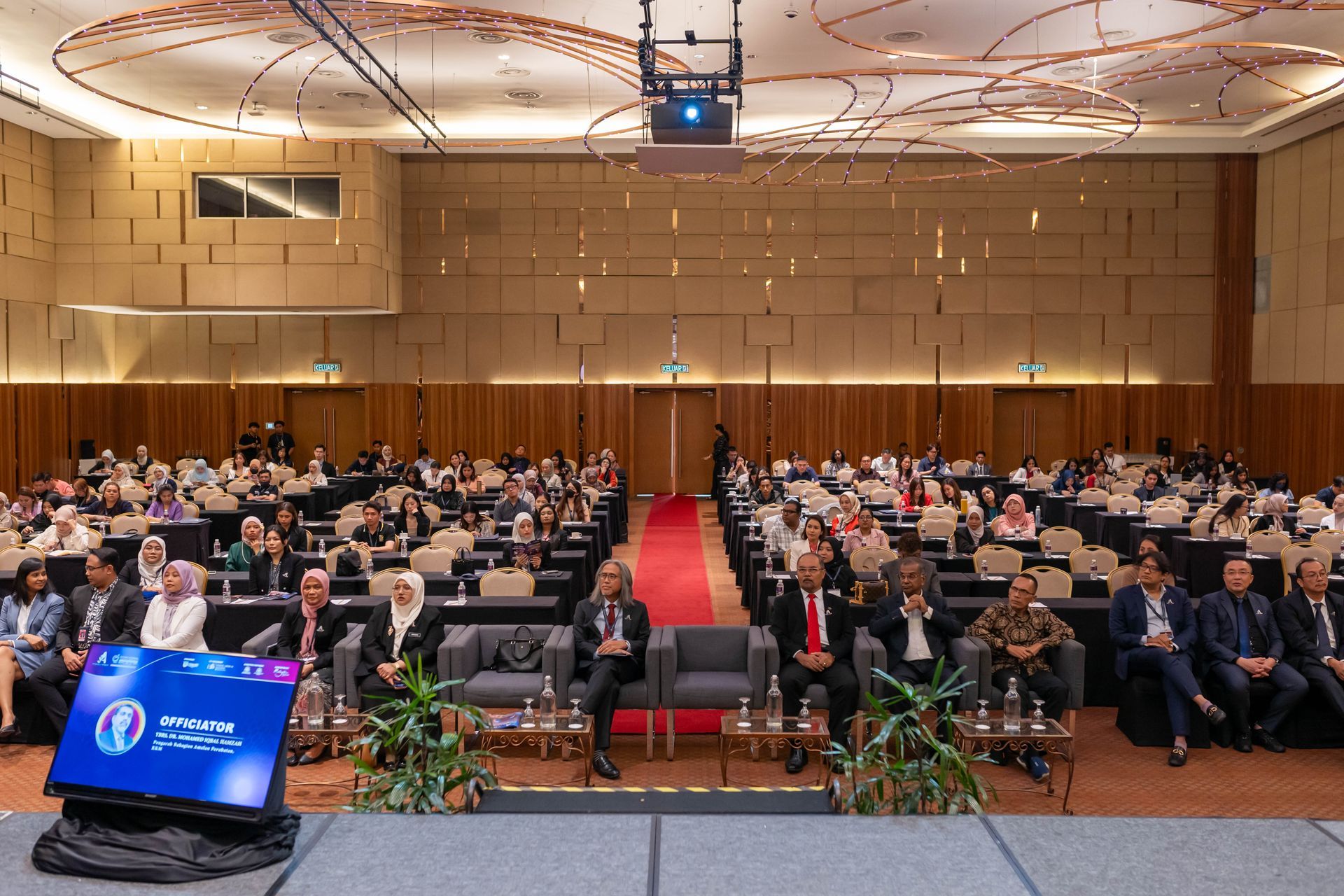 Conference hall with a panel seated on a stage, audience seated at tables, and a screen displaying text.