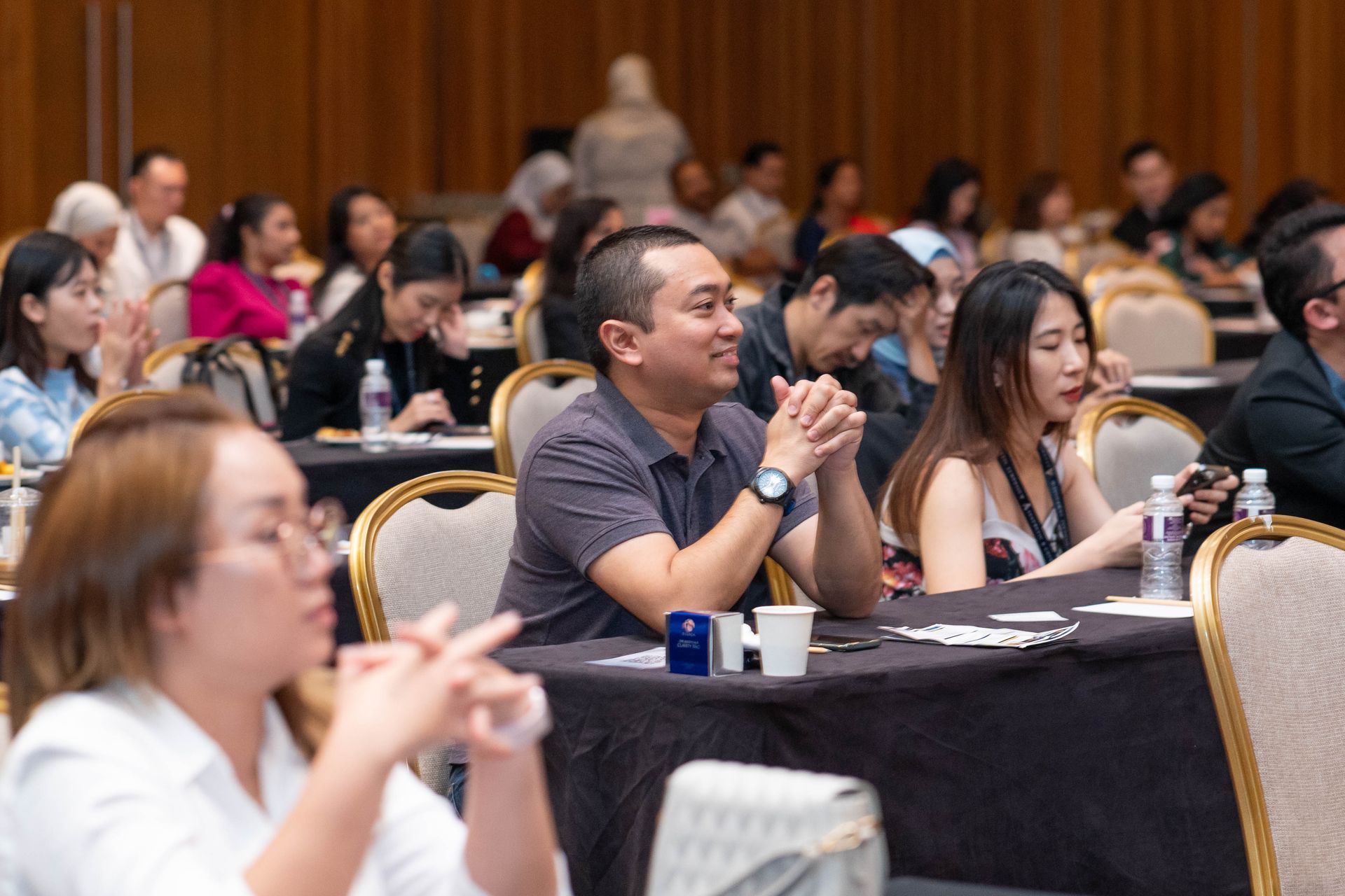 Attendees at a conference, some clapping, seated at tables with water bottles and name tags.