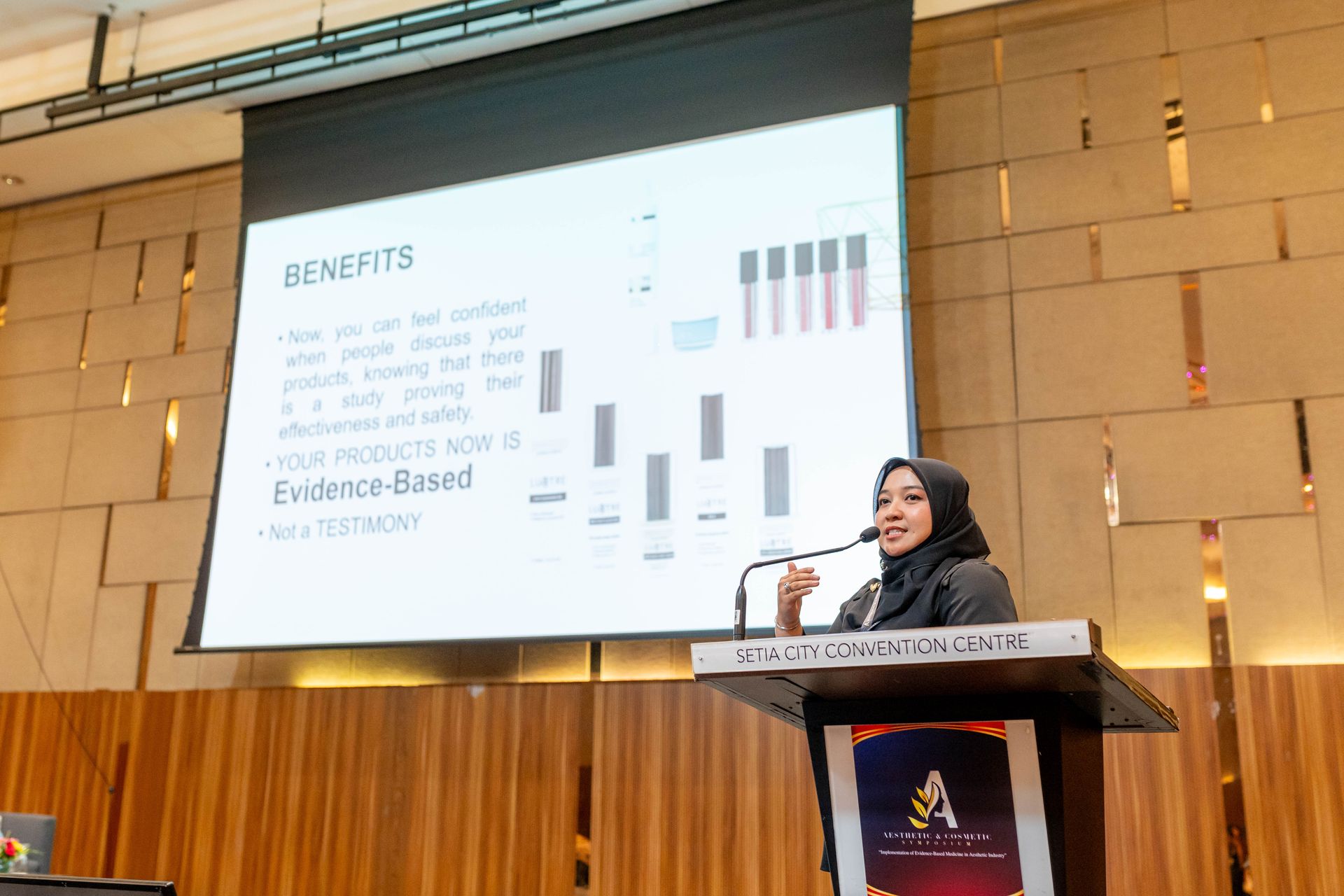 Woman in hijab giving a presentation at a podium with a screen showing benefits and graphs.