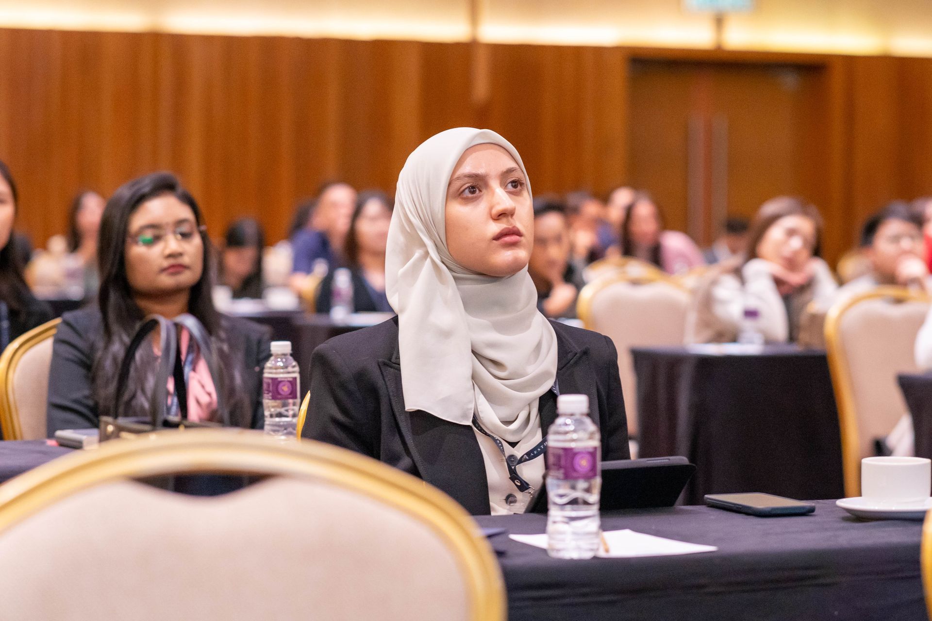 Woman in hijab at a conference, looking up intently, seated at a table. Others in audience.