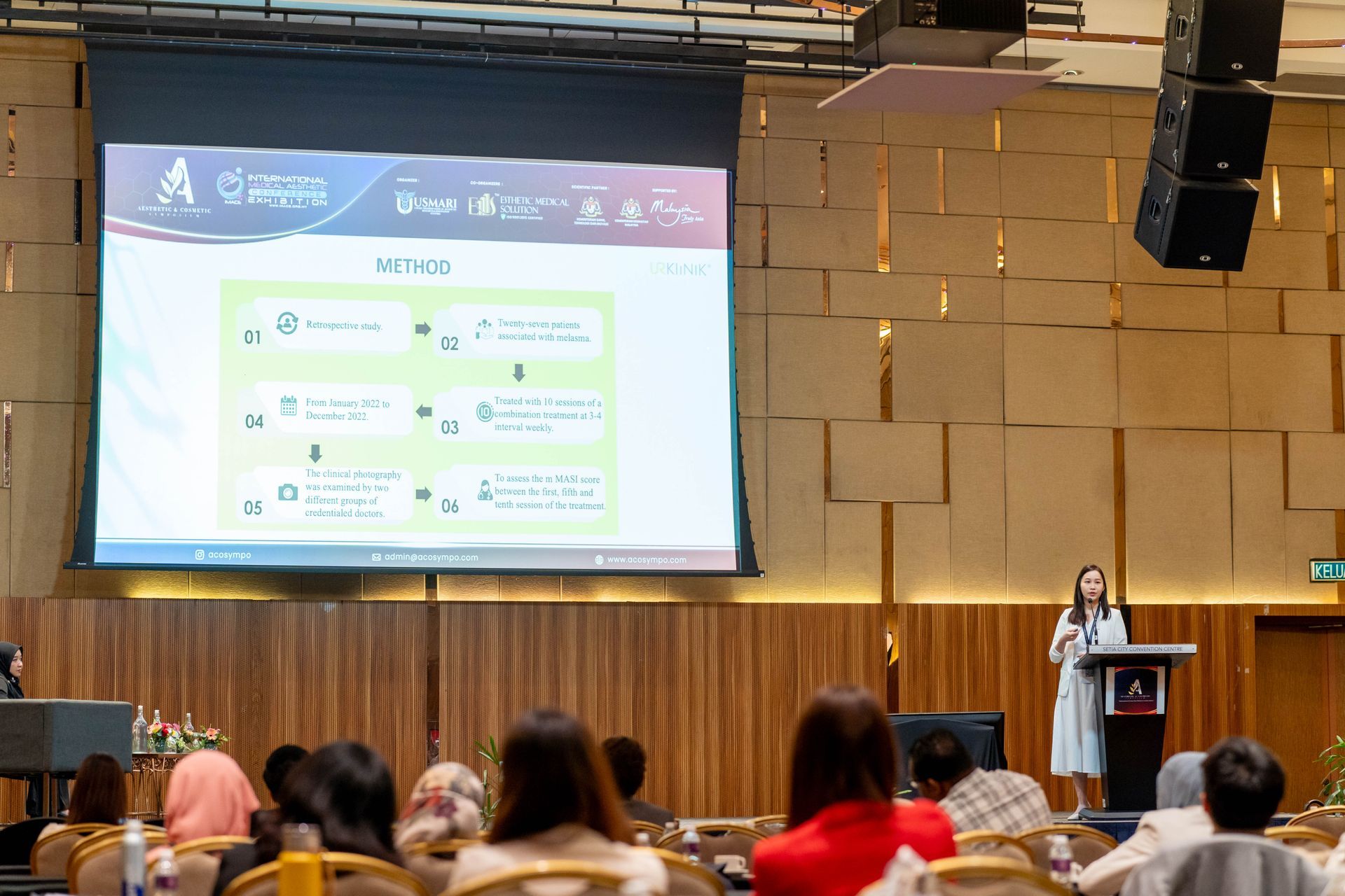 Woman presenting at a conference, large screen shows a diagram, audience seated.