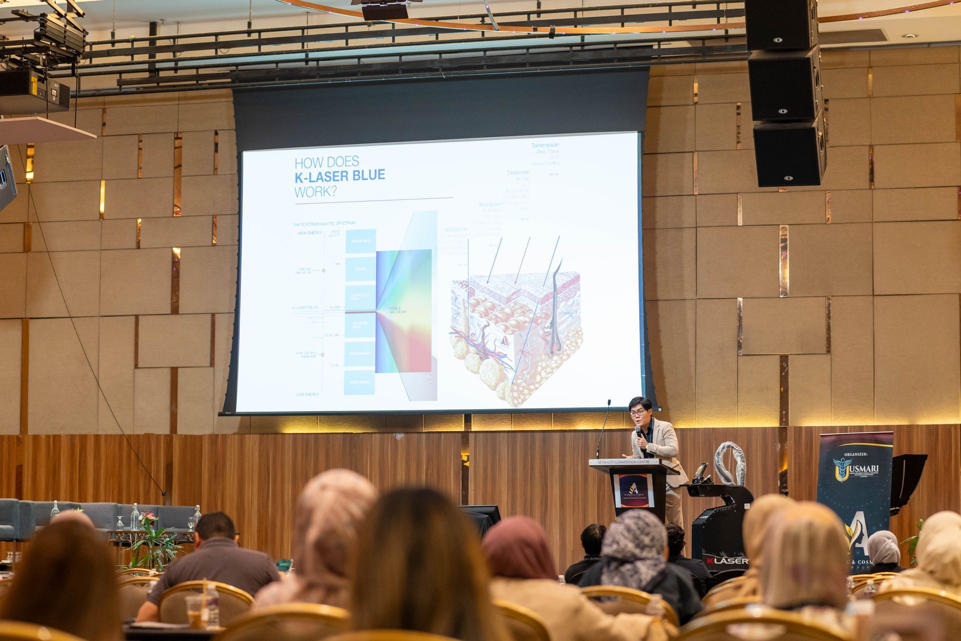 A person presents at a conference, with a slide showing a skin diagram and a color spectrum. The audience is seated.