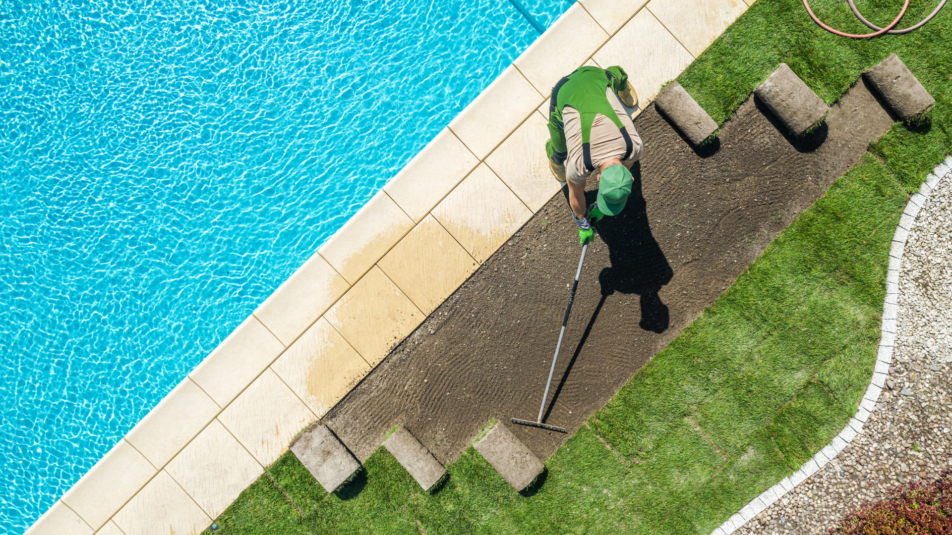 A man is raking the grass next to a swimming pool.