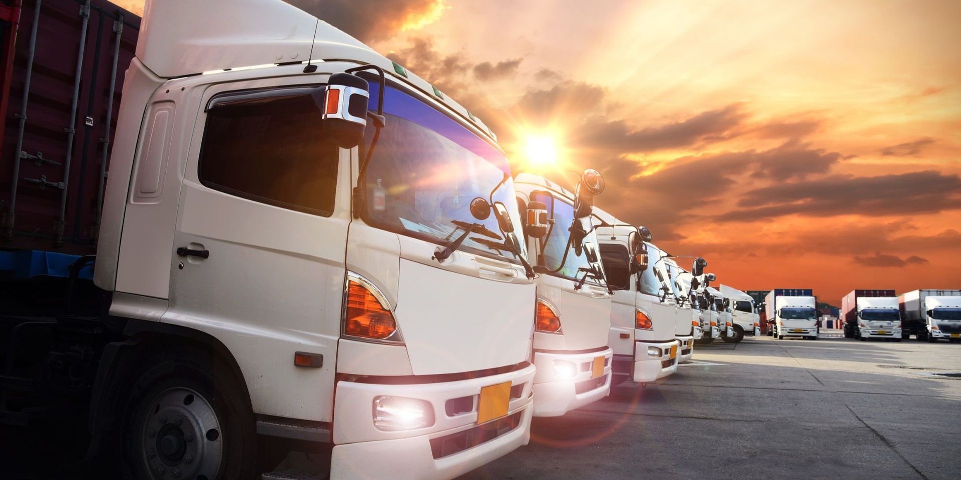 A row of semi trucks are parked in a parking lot at sunset.
