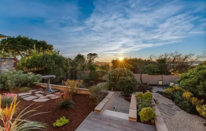 Sunlit garden courtyard with wooden deck, stone paths, shrubs, and trees at sunset